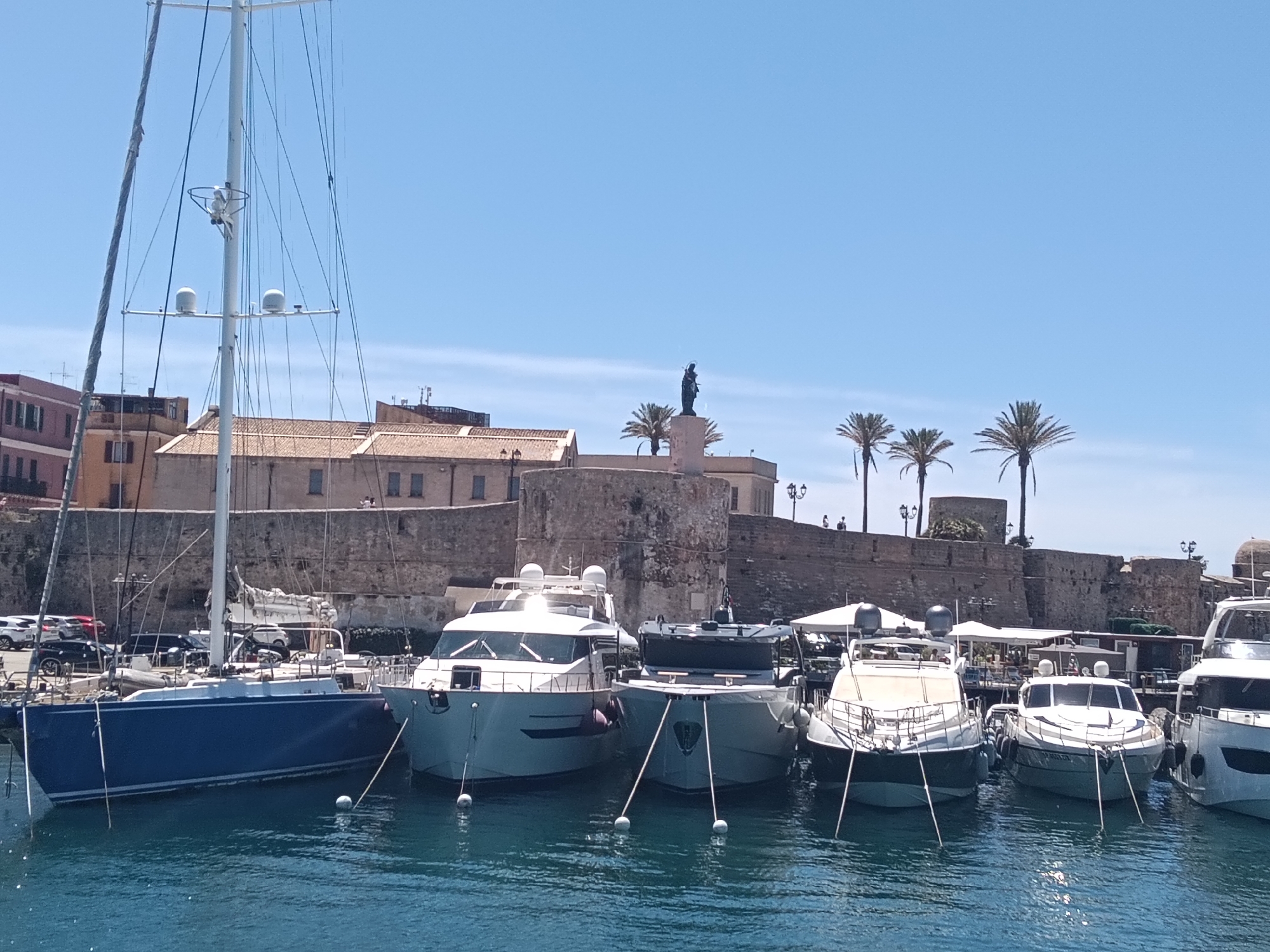 The image depicts a harbor scene in Alghero, Italy, with several yachts moored in the foreground. The yachts are of various sizes and designs, with clean lines and modern features. The water is a deep blue, reflecting the clear sky above. In the background, a stone wall, likely part of the city's fortifications, runs along the waterfront. Behind the wall, buildings with terracotta roofs are visible, suggesting an urban environment. Palm trees are scattered along the top of the wall, adding a tropical touch to the scene. A statue of a figure stands on a pedestal atop the wall, overlooking the harbor. The overall impression is one of a picturesque and tranquil setting, typical of a Mediterranean coastal town.