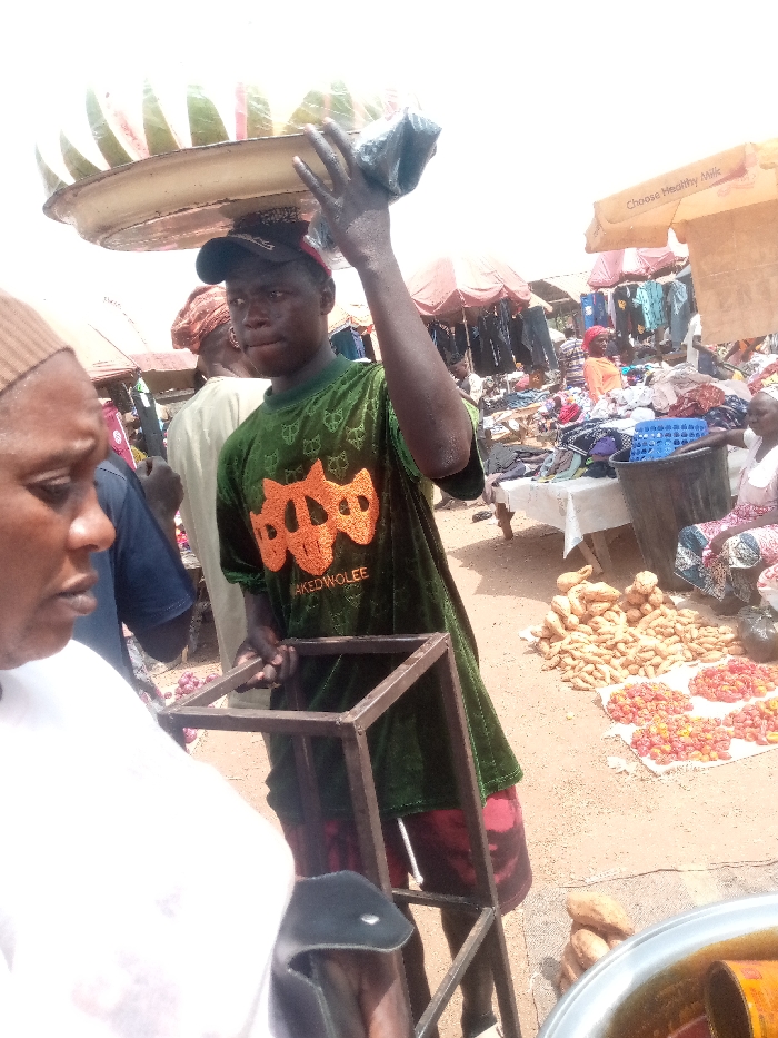The image depicts a bustling outdoor market scene during bright daylight, characterized by numerous vendors, goods, and people.
Central to the foreground is a young man, likely a street vendor, carrying a large, flat, light-colored round tray on his head. The tray is laden with evenly cut slices of what appear to be watermelon. He wears a dark baseball cap and a dark green, short-sleeved shirt that has a textured, possibly velvet, appearance. Prominent orange, abstract, skull-like designs are printed on the front of his shirt, above the visible text "NAKEDWOLEE". With both hands, he holds a dark metal square-framed object, which resembles a stand or a small stool. He has a focused expression, looking slightly to his left.
To the immediate left and partially cropped, the profile of another person, possibly female, is visible. This individual wears a white top and a brown head covering.
The background reveals the wider market environment. Various stalls are set up under colorful awnings and umbrellas, some red and others orange. One orange awning in the upper right corner displays the text "Choose Healthy Milk". Beneath these awnings, clothing items are seen hanging on rails and piled on tables, indicating clothing vendors. Several other people, including a person in an orange top and a woman sitting on the ground tending to goods, are visible in the mid-ground, contributing to the busy atmosphere.
On the ground to the right of the central figure, various produce items are displayed on white sheets. These include piles of light-colored, elongated vegetables, possibly sweet potatoes or yams, and several mounds of small, round, red items, which could be peppers or small tomatoes. A dark plastic bucket is also visible near these goods. In the bottom right corner, a portion of a large metal pot or bowl containing a liquid, possibly oil or a stew, is visible.
The ground throughout the market is unpaved, appearing as dirt or sand. The bright lighting suggests it is daytime, likely mid-day, with strong sunlight casting minimal shadows directly beneath objects due to the overhead perspective.