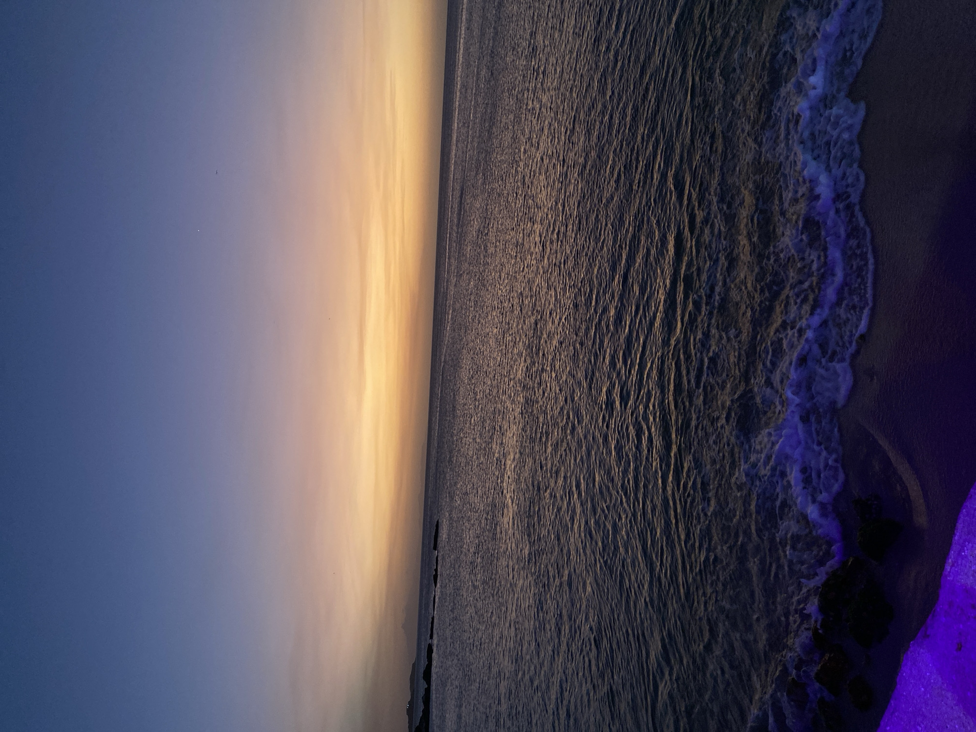 The image shows a beach scene at dusk or dawn. The sky transitions from a deep blue to a lighter yellow-orange hue near the horizon. The ocean spans the majority of the image, reflecting the colors of the sky. The water is dark with subtle ripples. Waves are visible at the shoreline, creating white foam as they break. Rocks are present near the edge of the beach. The location is likely Juan Griego, Venezuela, given the prompt.