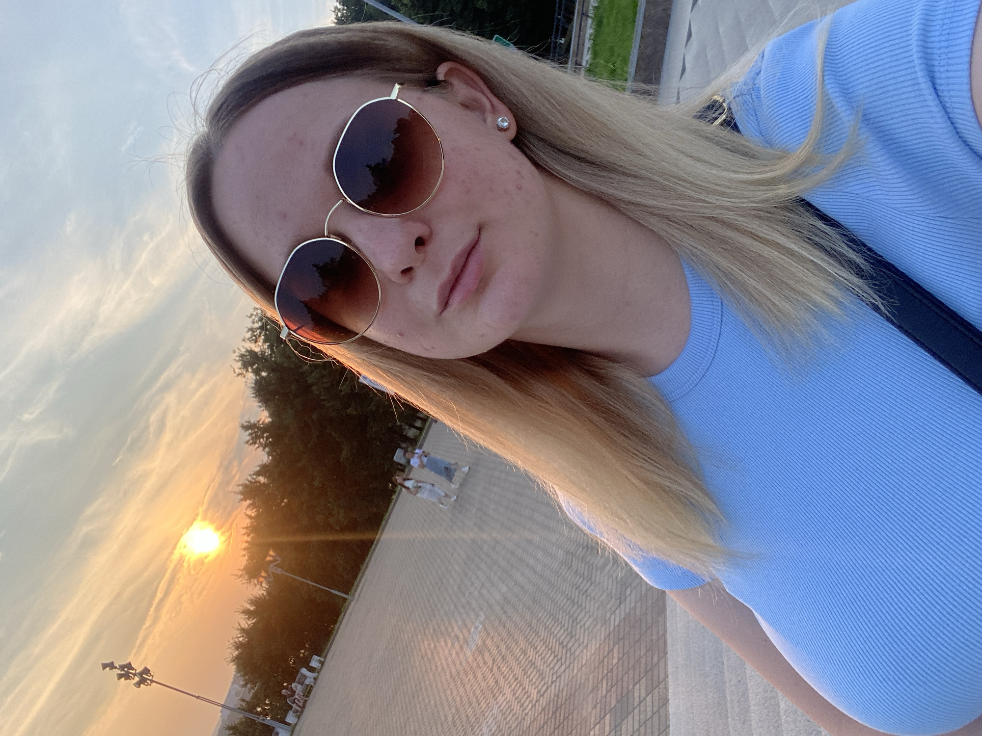 A young woman with blonde hair and sunglasses takes a selfie outdoors at sunset. She is wearing a light blue ribbed t-shirt and a small diamond stud earring. The background shows a paved area with trees and the sky. The sun is visible in the sky, casting a warm glow. The scene is likely set in Izhevsk, Russia, possibly in a park or public square.