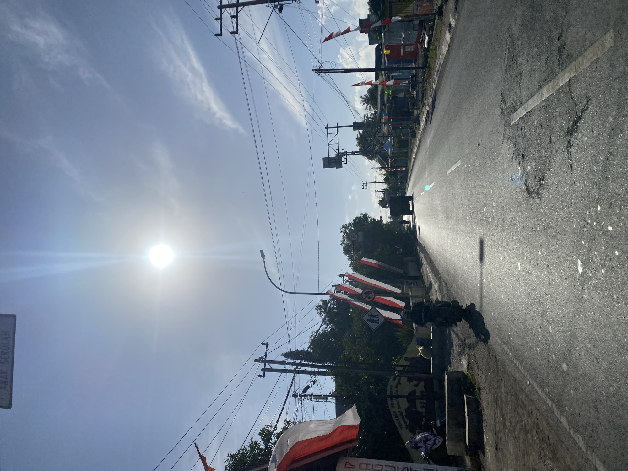 The image depicts a sun-drenched, paved road in Jalanjang, Indonesia, under a bright blue sky with scattered wispy clouds. The sun is positioned high in the sky, causing lens flare and casting strong shadows. The road itself shows signs of wear, with visible potholes and a white line marking the center.
To the left of the road, there is a grassy area with a low concrete barrier and a small ledge. On this ledge, a woman wearing a hijab is seated on a motorcycle. Next to her, another motorcycle, also ridden by a person wearing a dark helmet and jacket, is positioned on the road. Further back along the left side of the road, a banner with red and white stripes is visible, along with other red and white flags adorning poles. A sign with Indonesian text that translates to "AREA SEKOLAH" (School Area) is also present, as are traffic signs indicating a pedestrian crossing and a prohibition.
The right side of the road features several utility poles with numerous electrical wires crisscrossing overhead. Buildings with corrugated metal roofs and walls are situated along this side, with more red and white flags displayed. In the distance, a truck is seen driving away from the camera. The overall scene suggests a typical daytime setting in a somewhat rural or suburban area of Indonesia, possibly near a school, given the signage.