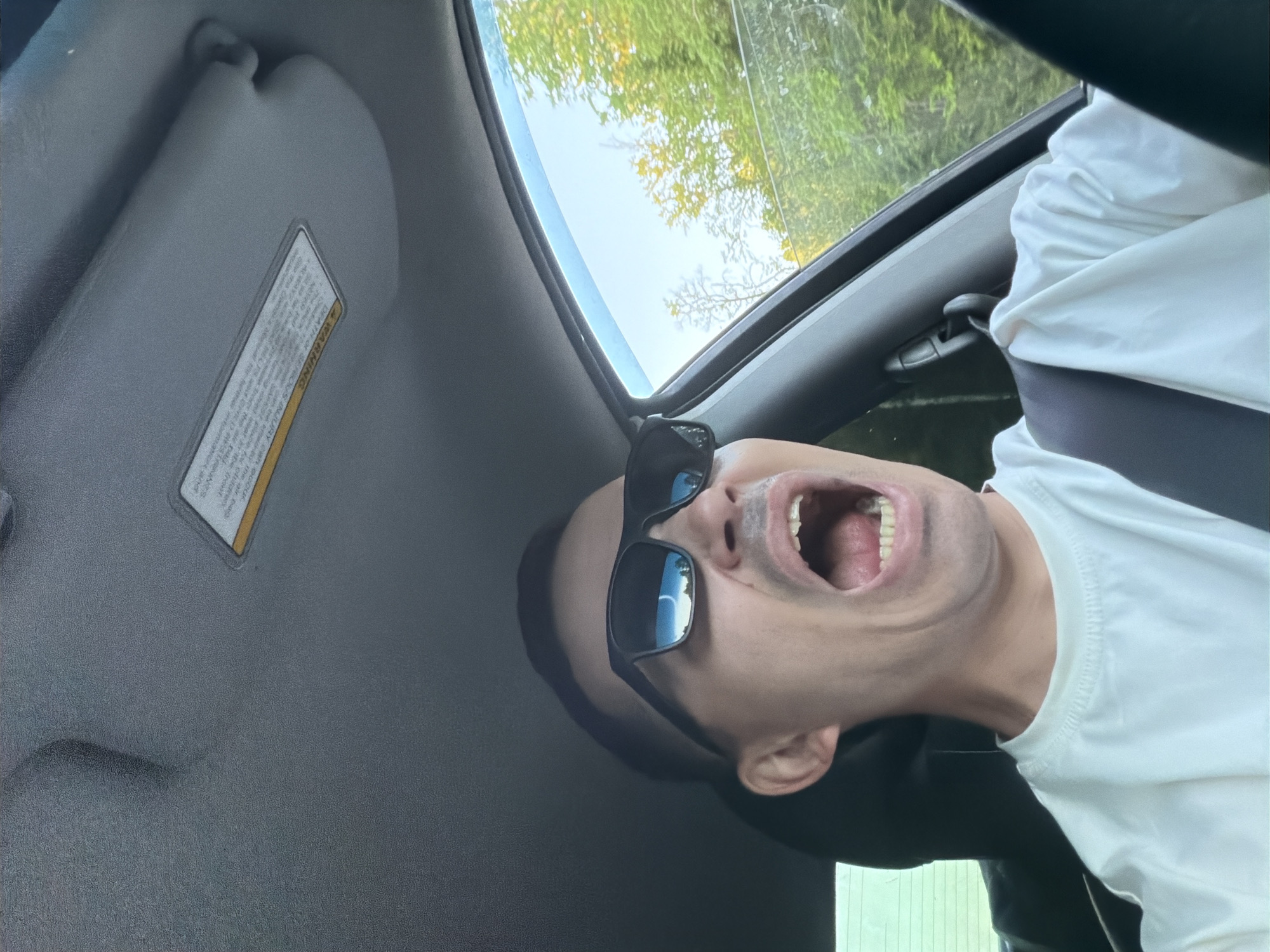 A man is sitting inside a car. He is wearing sunglasses and a white t-shirt. His mouth is open. The car is in an unknown location, with trees visible through the sunroof. The interior of the car is gray, with a sun visor on the left side. There is no information to indicate the location is in Severnyy, Russia.