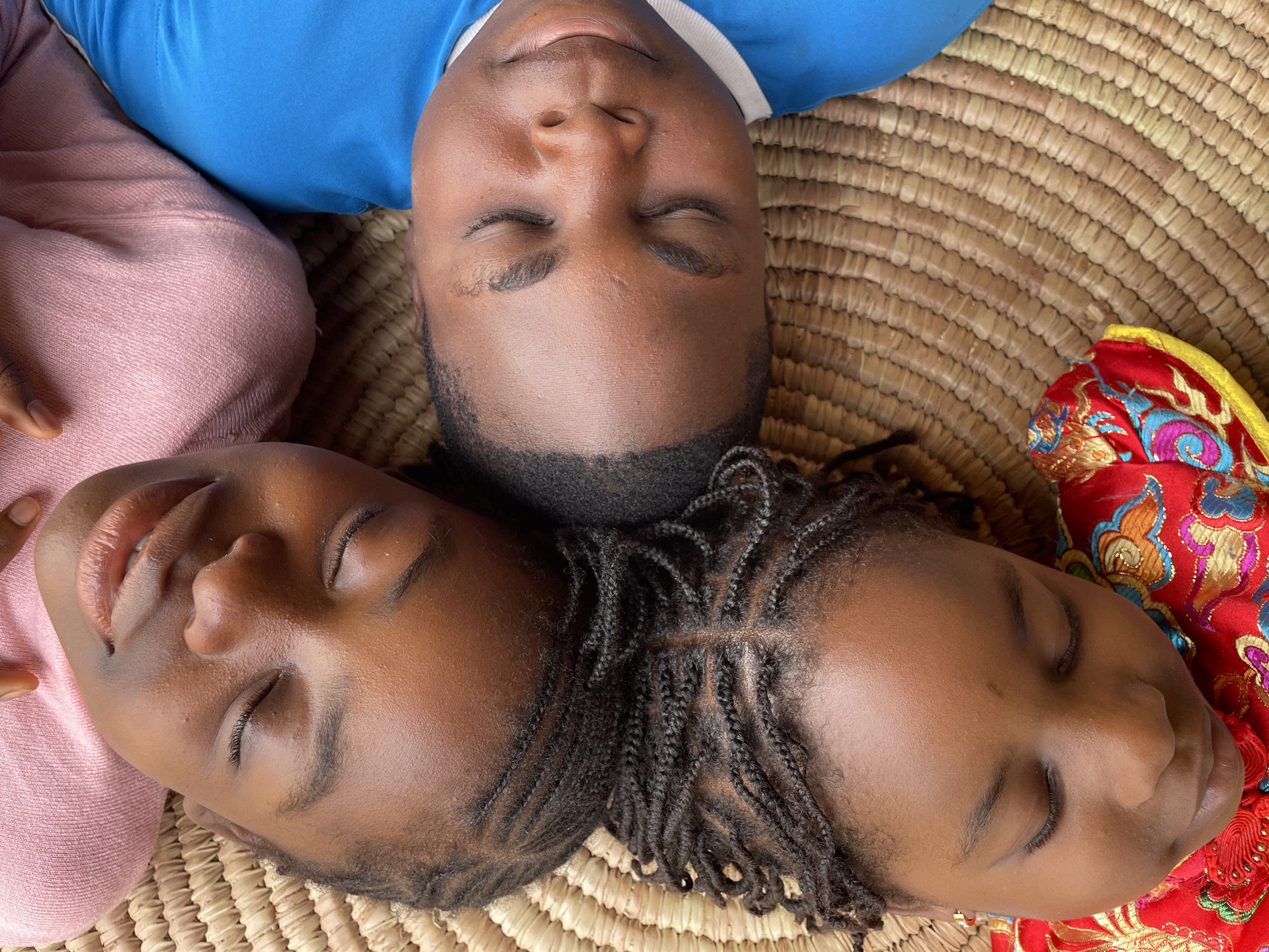 Three people are lying with their heads together on a woven mat. A person wearing a blue shirt is at the top of the frame. To the left of the person in blue is a person wearing a pink shirt. The person on the right is wearing a red and gold shirt. All three have their eyes closed.
