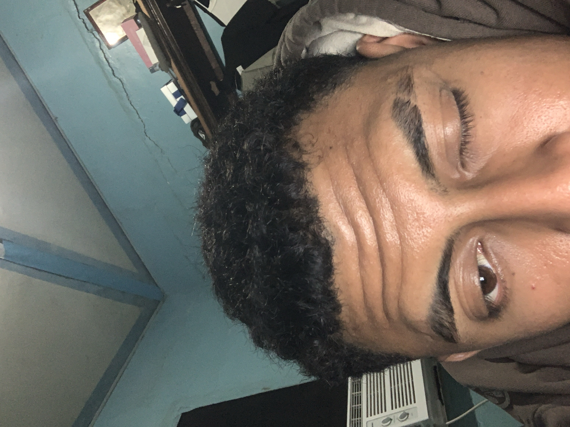 The image is a close-up, low-angle shot of a person's face, focusing on their forehead, eyes, and hair. The person has dark, curly hair and prominent forehead wrinkles. Their right eye is open and brown, while the left eye is partially closed. They appear to be wearing a brown hooded garment. The background consists of a light blue wall with a crack, a shelf with some items including boxes and a framed picture, and a portion of a window air conditioning unit. The overall lighting suggests an indoor setting, possibly at night or in a dimly lit room. The image does not provide any information that would allow for localization to Santa Ana, Venezuela.