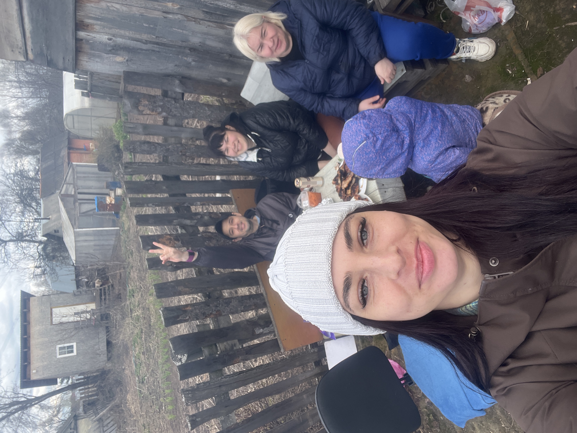 The image depicts a group of five people outdoors, seemingly in a rural or semi-rural setting.
People:
-A woman with dark hair and a white knitted hat is taking a selfie. She is wearing a brown jacket.
-Three other women are visible behind her, positioned around a table.
-A man is seated behind them, visible in the background, making a peace sign.
Objects:
-A wooden table is present, with various items on it including food and a bottle.
-A white plastic shopping bag sits near the group.
-Buildings, including one that appears to be made of weathered wood, are visible in the background.
-A black chair is visible.
Actions/Interactions:
-The woman with the hat is taking a selfie of herself and the group.
-The others are seated around the table. The mood appears relaxed.
-The man in the background is making a peace sign.
Environment:
-The scene appears to be in a backyard or outdoor area with a rustic feel.
-The ground appears to be covered in dirt and some sparse vegetation.
-The sky is overcast, suggesting cloudy weather.
Location:
-Chaikovsky, Russia. The location is likely an outdoor space within the city or on its outskirts, potentially a dacha or a rural residence. The structures and overall environment suggest a less urbanized area.