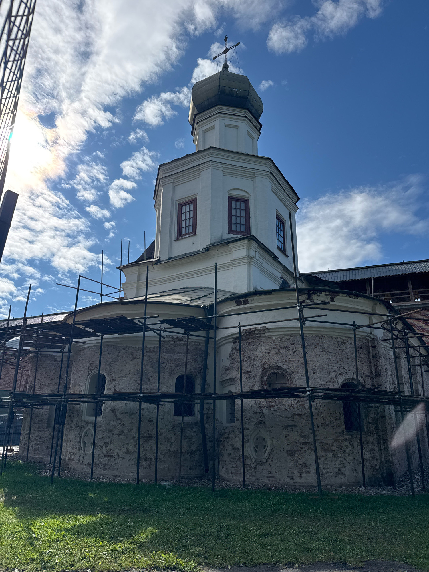 The image shows the St. George Cathedral of the Yuriev Monastery in Veliky Novgorod, Russia. The cathedral is partially covered in scaffolding, suggesting it is undergoing restoration or maintenance. The cathedral is constructed of stone and has a white upper section topped by a green dome with a cross. The lower section of the cathedral is made of rough stone with arched windows. A bright sun shines in the upper left corner, casting lens flare across the sky, which is blue with scattered white clouds. The foreground consists of a green lawn.