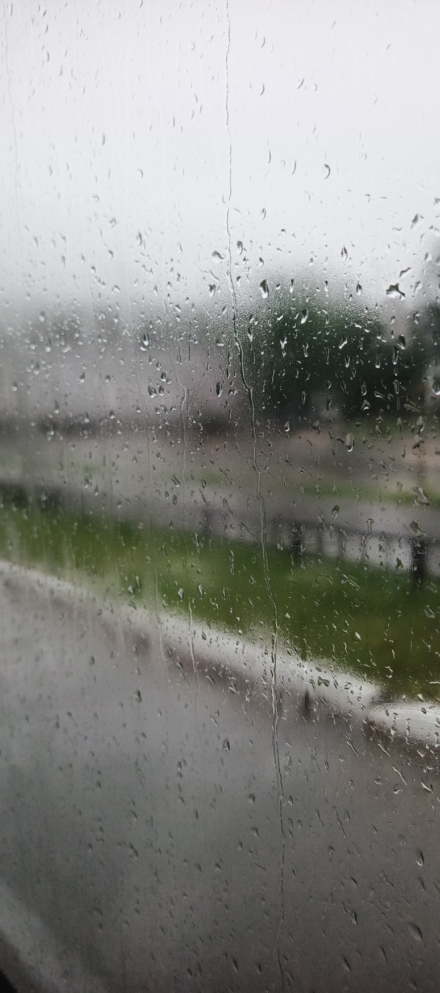 The image is a close-up, slightly out-of-focus shot of raindrops on a window. The raindrops create streaks and droplets on the glass, distorting the view of the outside world. Through the rain-streaked window, a blurred scene of what appears to be an urban or suburban environment can be seen. The background is dominated by muted colors of green and gray, suggesting foliage and buildings or infrastructure. A grey, wet road or pathway is visible in the mid-ground, separated from a green area by a dark railing or barrier. The sky appears overcast and bright, consistent with rainy weather. The overall impression is one of a gloomy, wet day viewed from indoors. The specific location in the city of Unknown, Unknown cannot be determined from the image.
