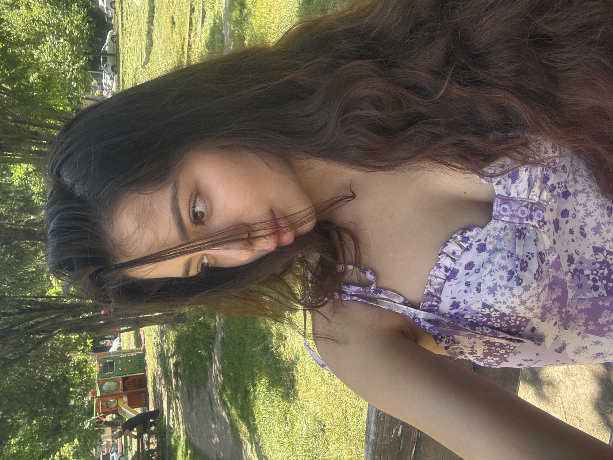 The image depicts a young woman with long, wavy, dark brown hair and light skin, wearing a purple and white floral-patterned dress. She is positioned outdoors, partially obscuring her face with her hair. The background includes a grassy area with trees, suggesting a park or garden. A playground structure is visible in the background. The location appears to be a park in an urban environment, possibly Plovdiv, Bulgaria. The woman appears to be seated, possibly on a bench, and looking towards the camera.