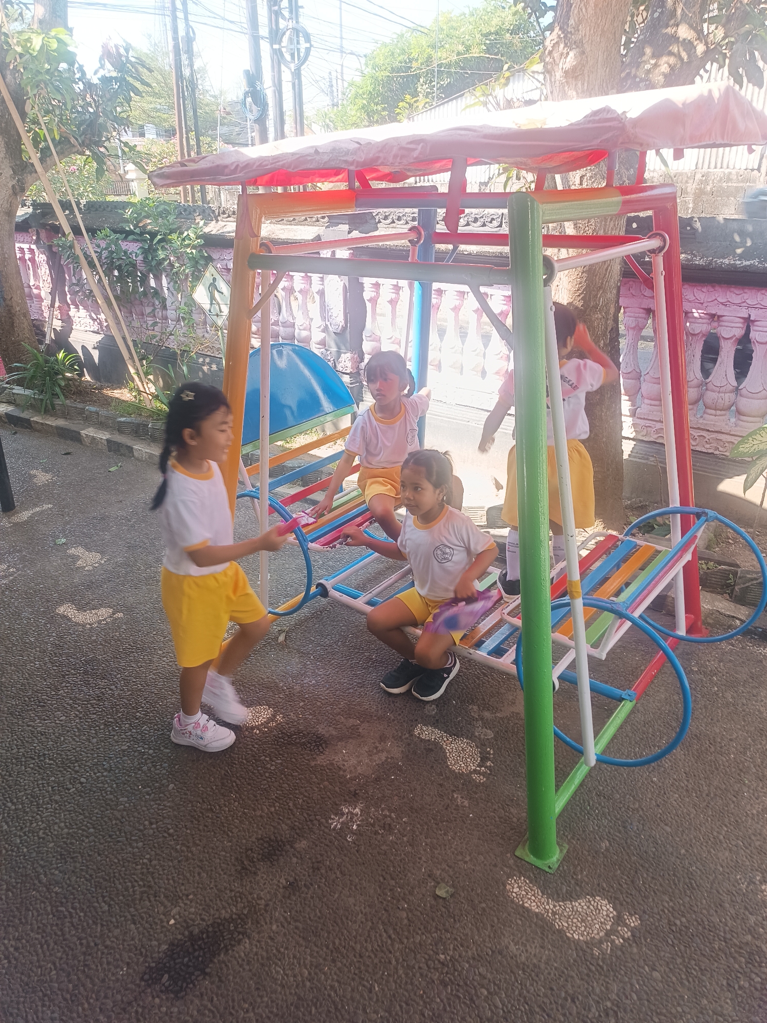 In Tabanan, Indonesia, a colorful playground swing set with a red awning is occupied by four young girls dressed in white shirts and yellow shorts. Two girls are seated on the colorful swing, while a third stands beside it, and the fourth is in motion on the left side of the frame. The ground is paved with small, dark pebbles, and a pair of white footprints are visible on the ground to the right of the swing set. The background features a tree, a pink-painted wall, and overhead utility poles and wires, suggesting an urban or semi-urban environment. The lighting indicates it is daytime, with shadows cast on the ground.
