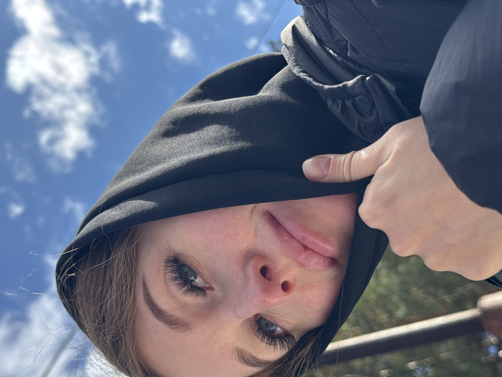 A close-up portrait shows a young person, likely female, looking directly into the camera from a low, inverted angle. Her head is tilted, and her face is partially framed by a black hoodie or jacket that she is wearing. Her left hand is visible on the right side of the frame, holding the fabric of the hood near her face.
The subject has fair skin, bright blue eyes with long, dark eyelashes, and neatly groomed eyebrows. Her lips are full and show a subtle, serene smile. A small stud piercing is visible on her left nostril. Light brown hair escapes from under the hood, framing her face.
The background features a clear, bright blue sky with scattered white clouds, suggesting a sunny daytime setting. Blurred green foliage and a dark, horizontal structure, possibly a railing or fence, are faintly visible in the lower right, indicating an outdoor location in Chaikovsky, Russia. The image has a casual and candid feel. No text is visible.