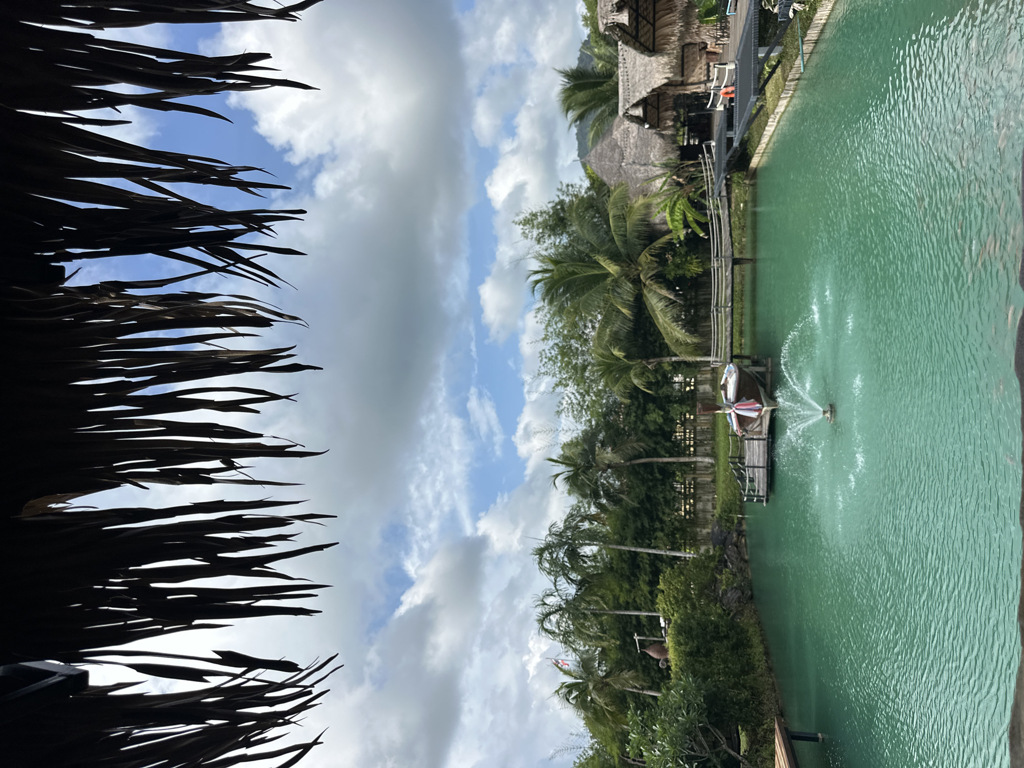 The image depicts a serene outdoor scene at Tambon Pa Kae Bo Hin, Thailand. In the foreground, a thatched roof overhang partially obscures the view of a vibrant turquoise body of water, likely a pond or small lake. A fountain sprays water in the center of the water. A wooden pier extends from the left side of the pond, and another wooden walkway with railings connects to a small boat docked on the right side. The boat is adorned with colorful fabric streamers. Beyond the water, lush green vegetation, including palm trees and dense foliage, forms a backdrop. In the distance, several traditional thatched-roof huts with conical roofs are visible, suggesting a resort or cultural establishment. The sky is partly cloudy with patches of blue, indicating fair weather. The overall impression is one of a tranquil tropical environment.