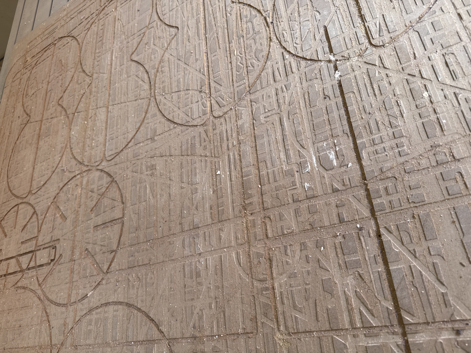 The image depicts an intricately carved wooden surface showing the street grid of the city of San Andrés Cholula, Mexico. The grid pattern is meticulously etched, with smaller rectangular shapes representing building blocks. Superimposed on the street grid are larger, curved shapes, some resembling clock faces with lines indicating the position of the clock arms. The wood surface has a dusty appearance with small white flecks scattered across it. The carving is detailed and precise.