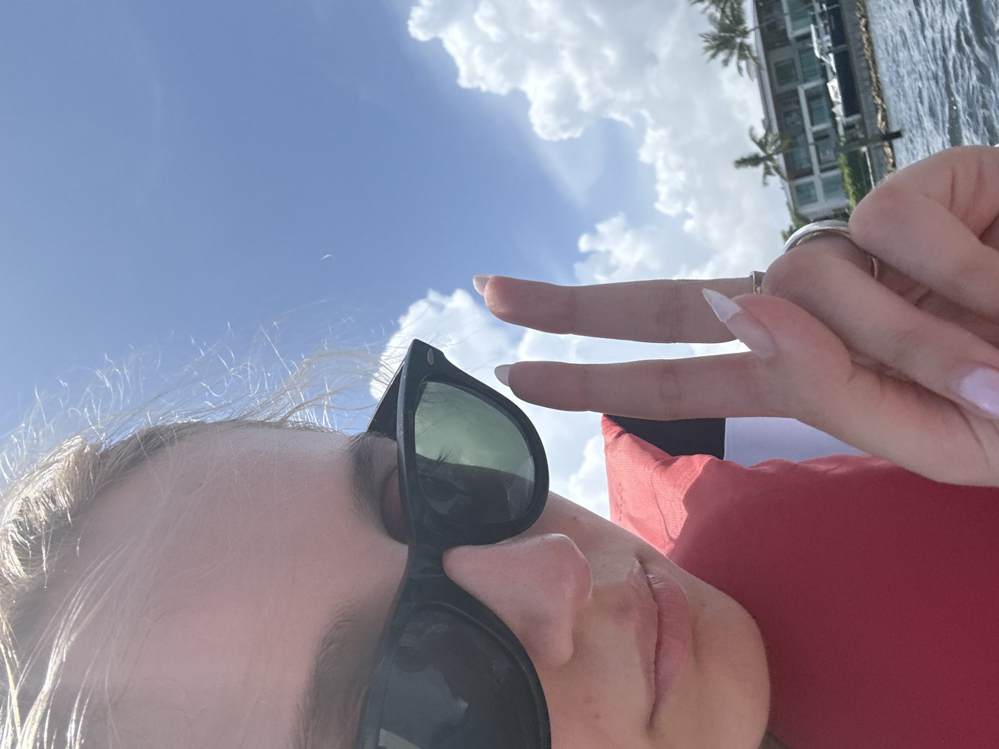 This media file is a selfie of a person outdoors on a sunny day. The person is wearing sunglasses and a red top. Their hand is in the frame, making a peace sign. In the background, there is a body of water, buildings with palm trees, and a cloudy blue sky. The image appears to be taken in Miami, Florida. The lighting suggests it is daytime, and the clear sky and sun indicate good weather. The overall mood is relaxed and cheerful. There is no visible text in the image.