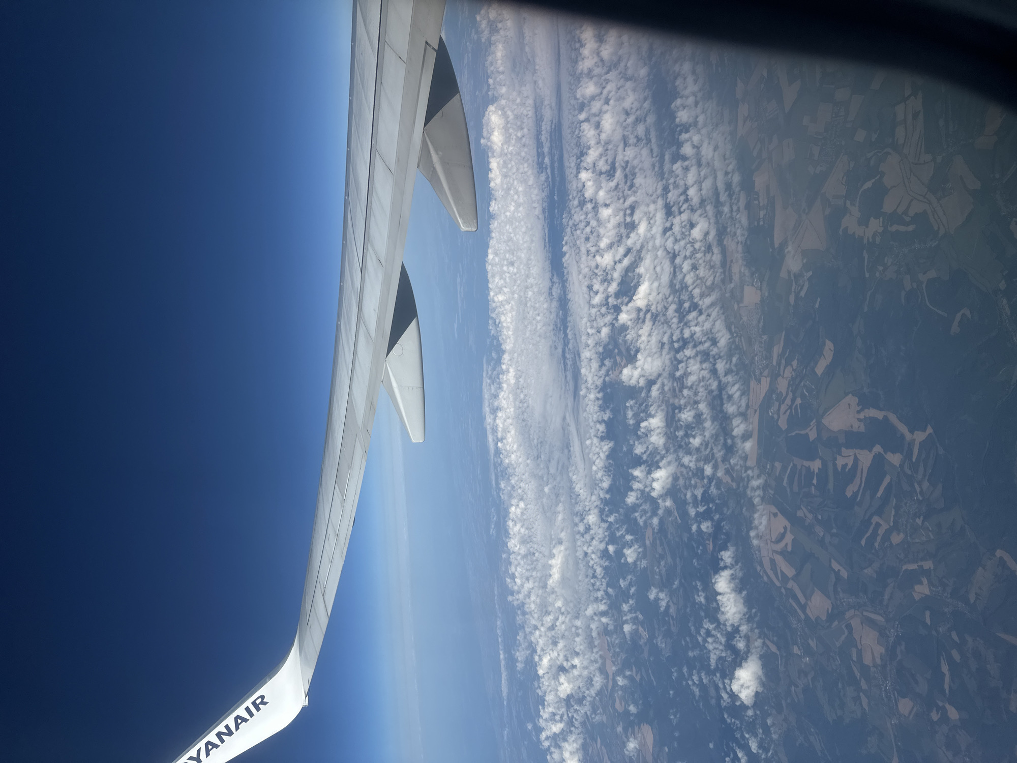 An aerial view from an airplane window shows the wing of the aircraft with the "RYANAIR" logo visible on its white surface. The wing extends across the upper right portion of the image, angled downwards. Below the wing, a vast expanse of the earth's surface is visible, covered by scattered white clouds and patterned land formations. The land appears to be a patchwork of fields with varying colors and textures, suggesting agricultural use, possibly in the vicinity of Tillé, France, given the context of a Ryanair flight. The sky above is a clear, deep blue.