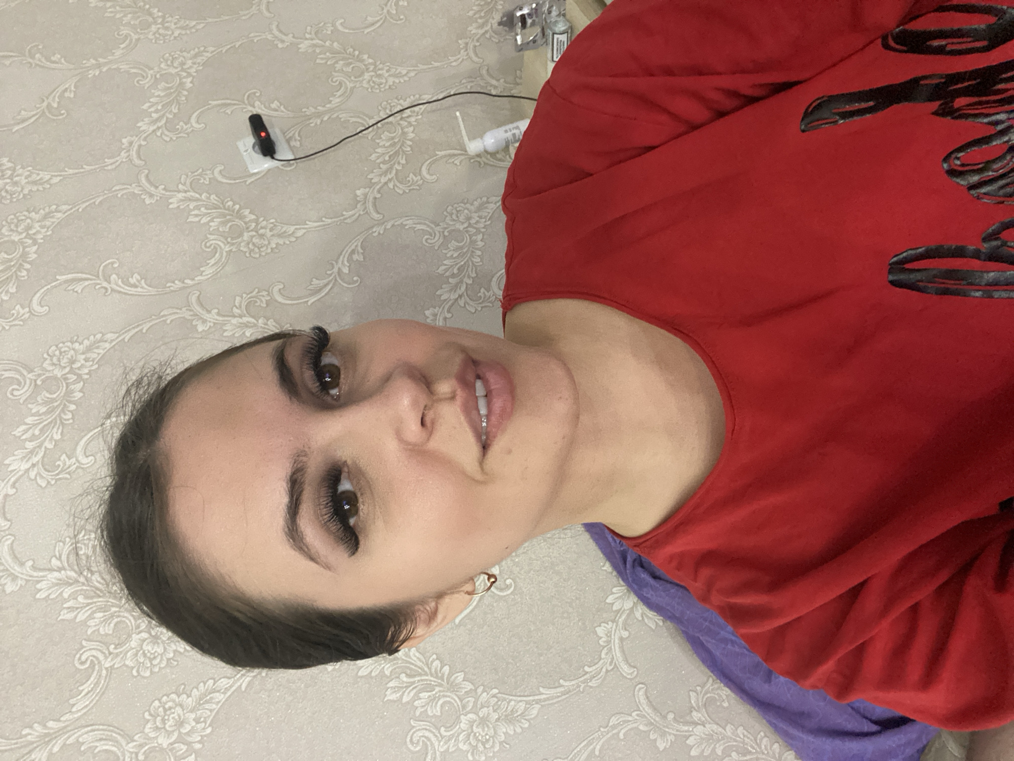 A woman is facing the camera, wearing a red t-shirt with black writing and a purple scarf/cloth. She has brown hair and eyelashes. The background features patterned wallpaper, an electrical outlet with a device plugged in, and a bottle. The location is likely indoors, potentially in Chaikovsky, Russia.