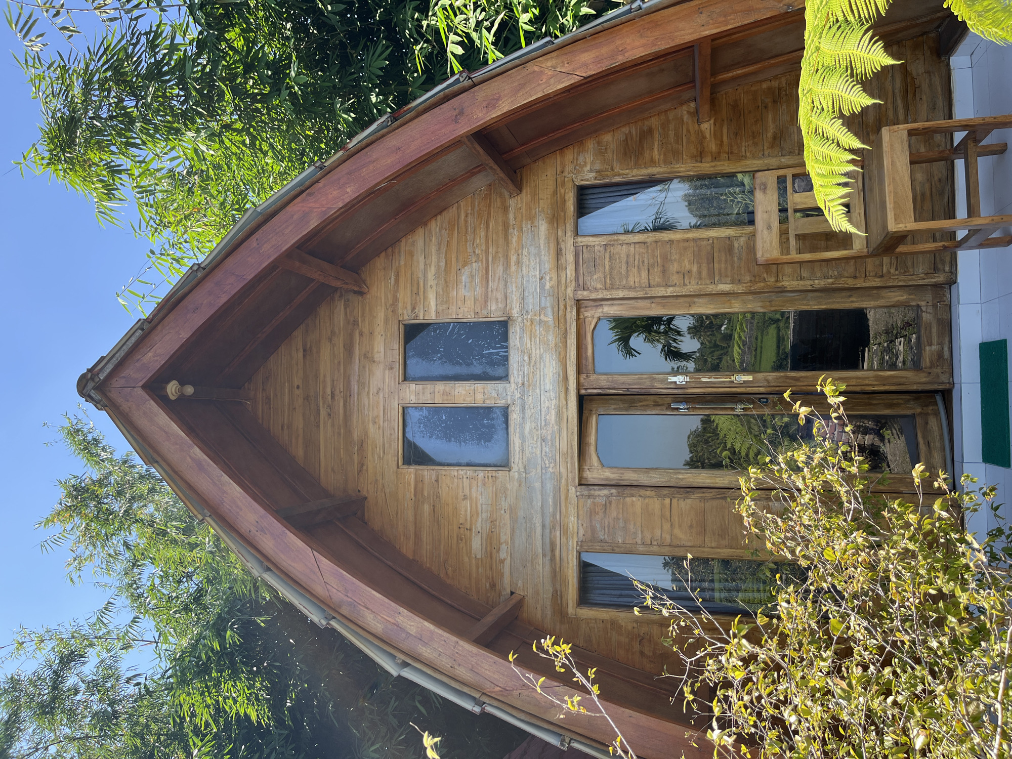 A wooden triangular-shaped cabin is set against a backdrop of lush green trees and a clear blue sky. The cabin features a large wooden door with glass panels and two smaller rectangular windows above it. To the right of the door, a simple wooden chair sits on a white tiled porch, partially obscured by a vibrant green fern. The cabin appears to be located in a tropical environment, possibly a resort or a homestay, given the surrounding vegetation and the outdoor seating. The overall scene suggests a tranquil setting in Tetebatu, Indonesia, a region known for its natural beauty and rice terraces.