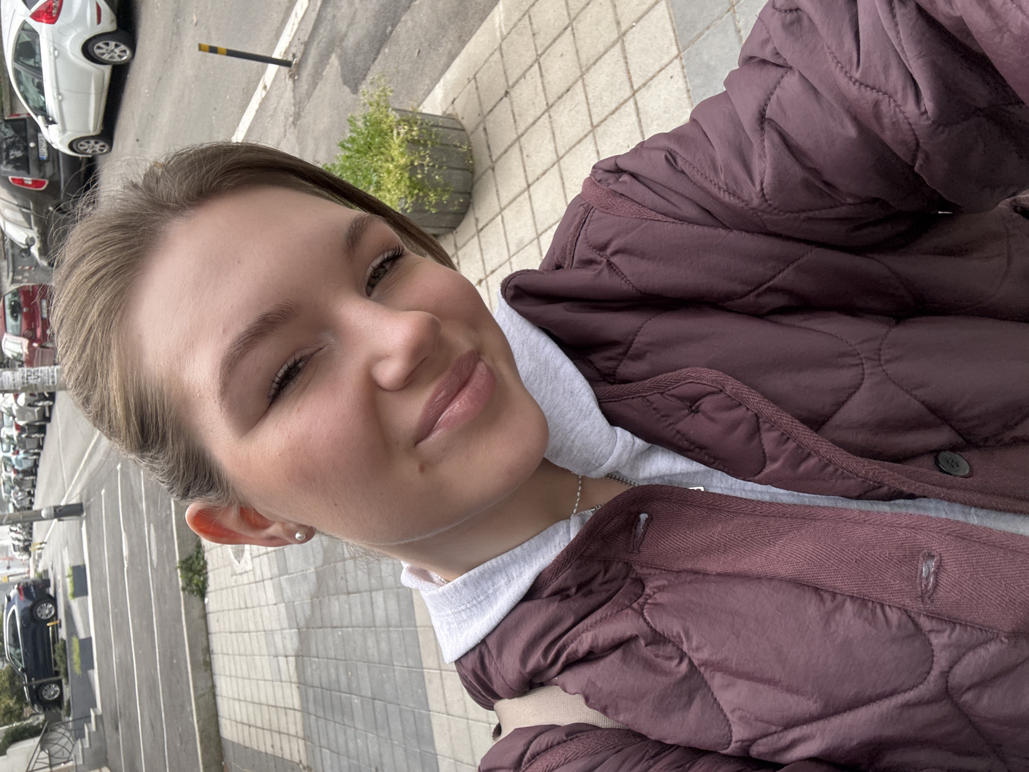 A selfie taken outdoors in Vozdovac Urban Municipality, Serbia, shows a young woman wearing a maroon quilted jacket over a grey hooded sweatshirt. She is standing on a paved area, likely a sidewalk, with a row of parked cars and buildings visible in the background. A planter with greenery is also visible. The lighting suggests it is daytime.