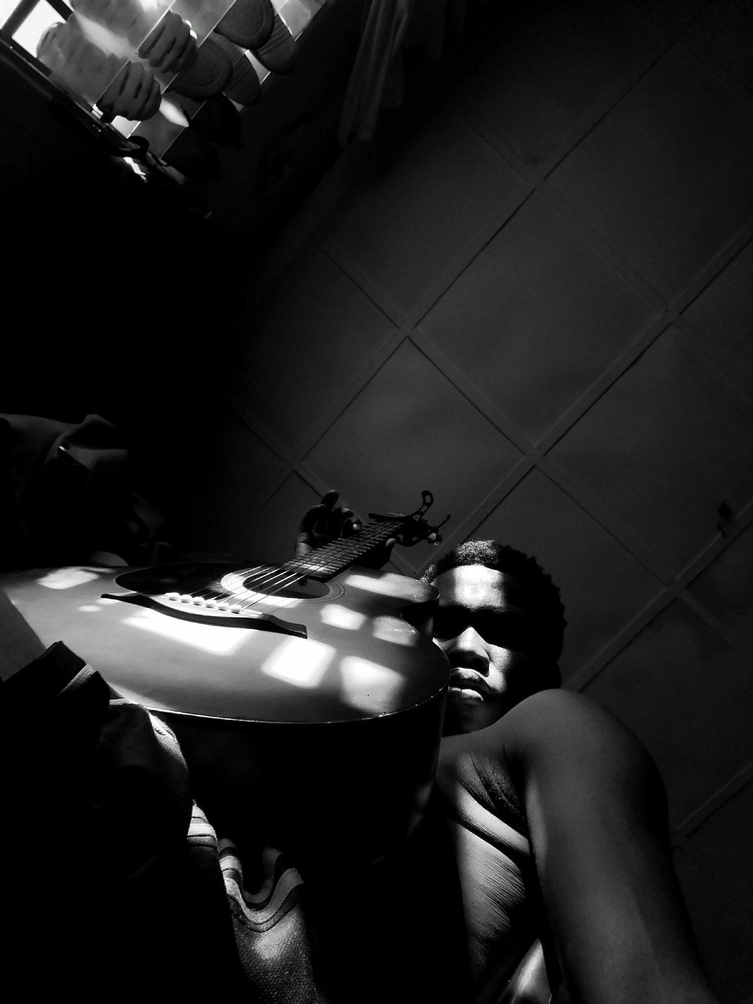 A low-angle, black-and-white photograph shows a dark-skinned person holding a guitar in an interior space in Keffi, Nigeria. The person is partially visible; the face is mostly shadowed, but a portion of the right shoulder and arm are illuminated. The guitar, positioned prominently in the foreground, is partially lit by light filtering through what appears to be a window with blinds, creating a pattern of light and shadow on its surface. A wall with square or rectangular panels is visible in the background. A window with blinds is also visible at the top left corner. The lighting is high contrast, creating stark shadows and highlights.