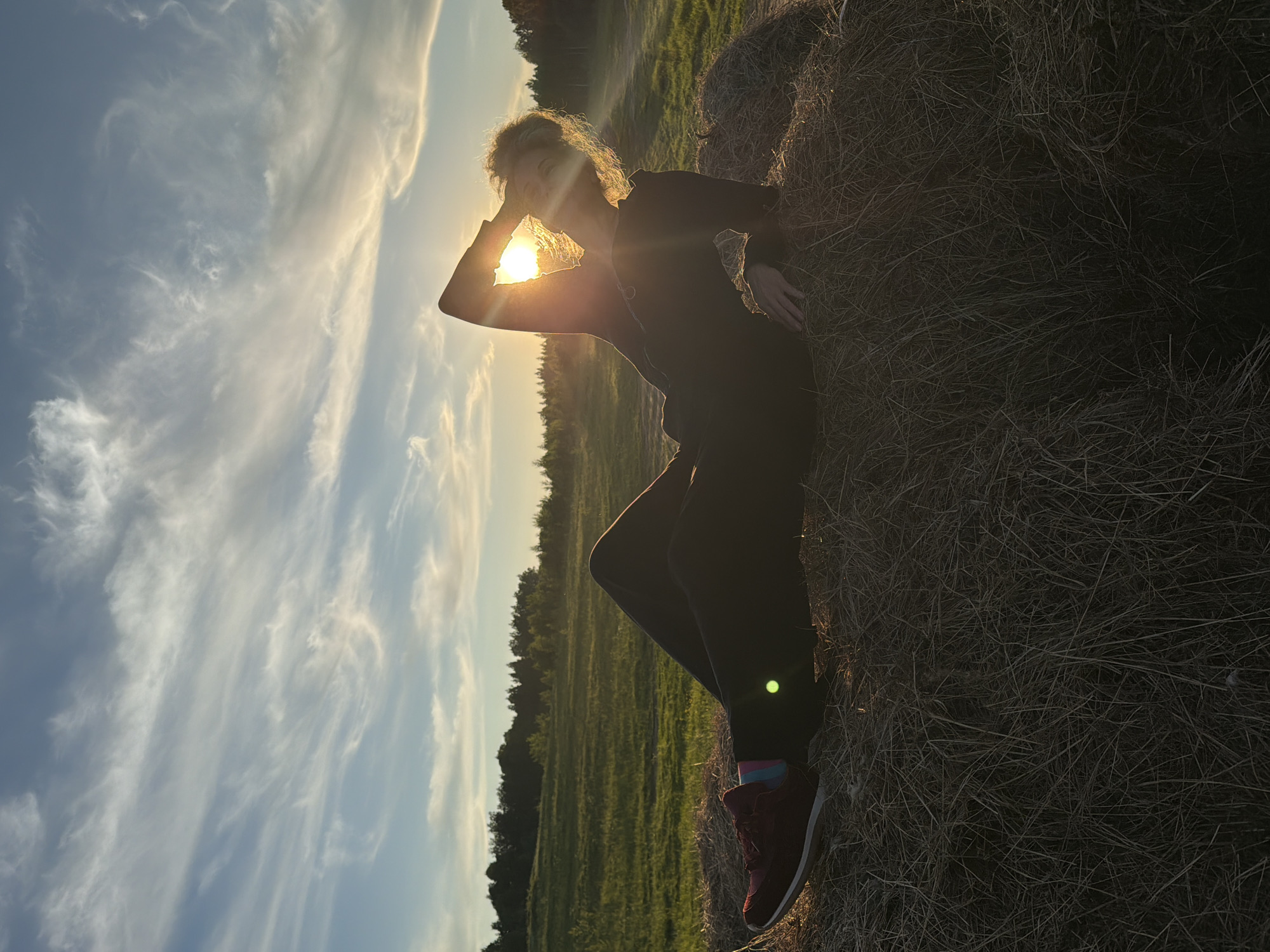 This image, set in Ozernoye-1, Russia, features a solitary figure silhouetted against a radiant sunset or sunrise in a natural, rural landscape. The image is presented in a vertical orientation but appears rotated counter-clockwise.
The central subject is a person, likely a woman with bright, curly hair, whose form is largely darkened by the strong backlighting. She is dressed in dark, presumably athletic, clothing consisting of a long-sleeved top and pants. Her footwear consists of distinct red sneakers paired with colorful socks. She adopts a posed posture, leaning her left side against a large, roughly textured hay bale positioned on the right side of the frame. Her left arm is bent, with her hand touching her forehead as if shading her eyes or in a thoughtful gesture, while her right hand rests on the hay bale.
The sun, a brilliant disc, is positioned behind and slightly above her head, creating an intense halo of light around her hair and body. This backlighting results in a dramatic silhouette effect and causes a visible lens flare near her left leg.
The background unfolds into a wide, verdant field or meadow, stretching towards a distant line of dark trees that mark the horizon. The sky above is painted with soft hues of blue and orange around the sun, fading into a lighter blue adorned with delicate, elongated white clouds, suggesting clear and serene weather conditions. The overall mood is peaceful and evocative of nature's beauty during the golden hour. There is no visible text in the image.