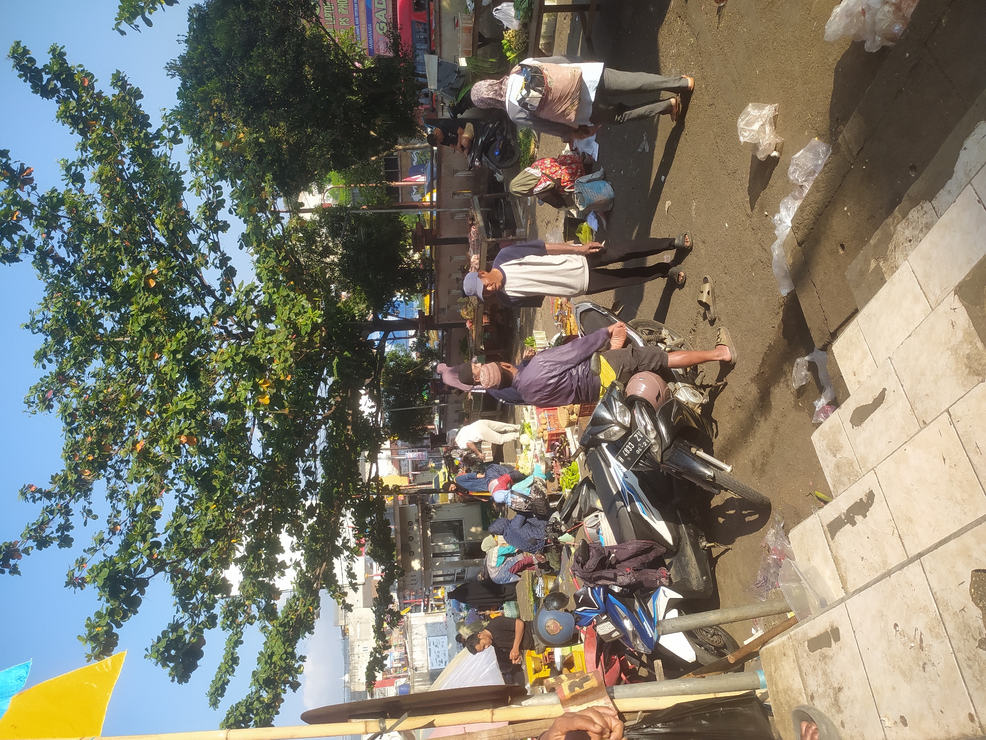 The image depicts an outdoor market scene in Kupang, Indonesia. The market is bustling with activity, with numerous people engaged in buying and selling various goods. Stalls are set up along a street, displaying produce, meats, and other items. Several motorcycles are parked, indicating a common mode of transportation in the area. The overall atmosphere suggests a vibrant and active marketplace.