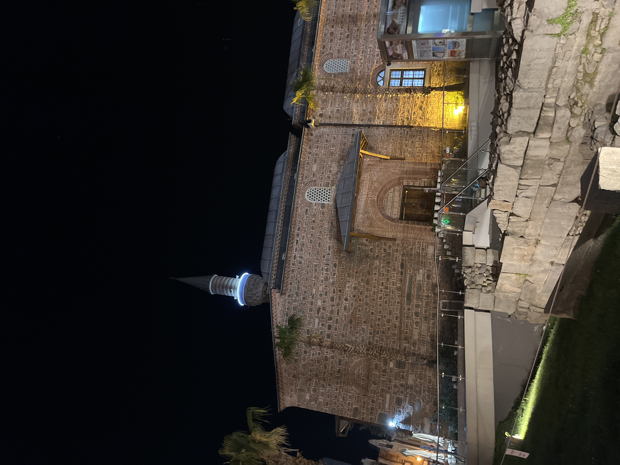 The image depicts the Dzhumaya Mosque at night, located in Plovdiv, Bulgaria. The mosque's minaret is visible on the left, illuminated with blue light. The main structure of the mosque is brick, with arched windows and a tiled roof. Adjacent to the mosque is a stone wall, and there is green foliage visible on the right side of the frame. The night sky is dark.