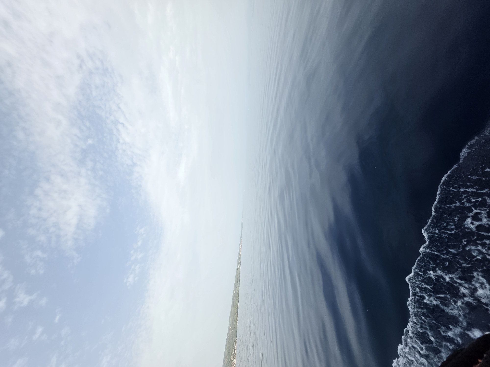 The image captures a serene seascape with a distant coastline under a pale, cloudy sky. The foreground is dominated by dark blue water with gentle ripples, transitioning into a lighter, hazy blue towards the horizon. On the left side, a white foamy wake, indicative of a moving vessel, cuts across the frame. The distant land appears as a muted green and brown strip, dotted with what seem to be small structures, likely the town of Postira on the island of Brač, Croatia. The overall atmosphere is tranquil and expansive, with a sense of calm at sea.