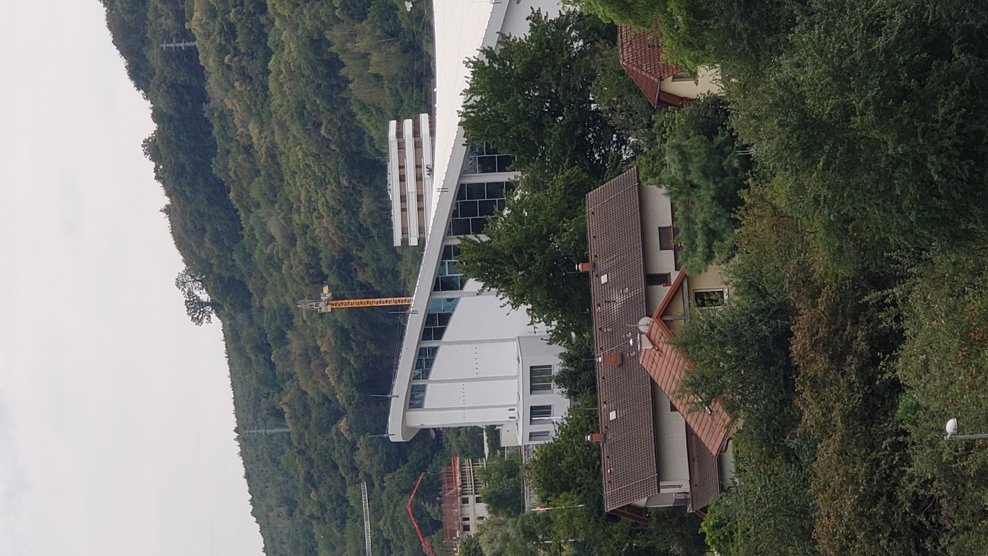 The image captures a panoramic view of a hillside in Prešov, Slovakia, dominated by lush green trees and vegetation. In the foreground, several residential houses with tiled roofs are visible. Beyond these homes, a modern white building with a distinctive angled roof and large windows stands out. A yellow construction crane is positioned on the hillside behind this building, suggesting ongoing development or renovation. Further up the hill, a taller, multi-story building with a stacked architectural design is present. The sky is overcast, contributing to a muted lighting condition across the scene. The environment appears to be a semi-urban or suburban area situated at the foot of a forested hill. There are no people visible in the image.
