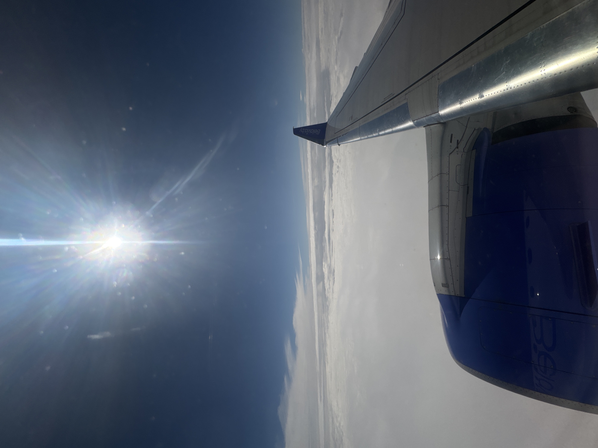 The image depicts an airplane wing and engine viewed from an airplane window, with a blue sky and sun visible outside. The wing has the logo "Belavia.by" printed on its tip. The airplane is flying above a layer of clouds. This image could have been taken while flying over Драчкаўскі сельскі Савет, Belarus.