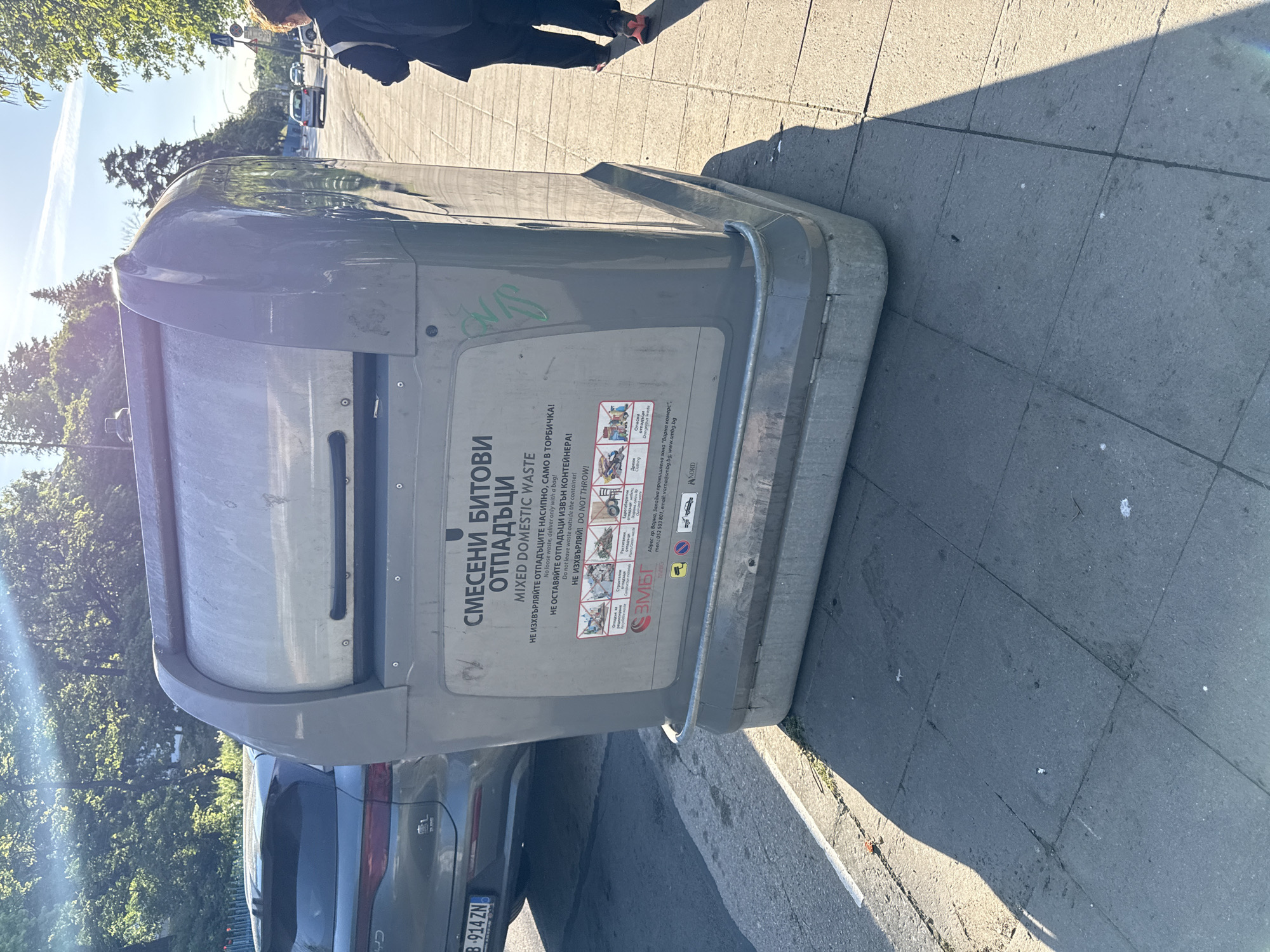The image shows a gray, metal mixed domestic waste container on a sidewalk, likely in a city. The container has Bulgarian and English instructions on its side. A car with license plate B-914 ZN is visible behind the container. A person wearing dark clothing is walking away in the background. The sidewalk is made of gray rectangular stones. Trees are visible in the background. The lighting suggests it's daytime.