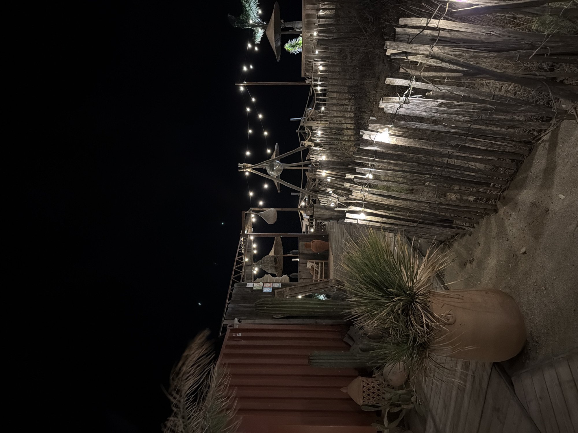 The image depicts an outdoor area at night, possibly a restaurant or bar, located in Torreilles, France. The scene is illuminated by strings of lights and a disco ball suspended from a wooden structure. A rough, wooden fence runs along the foreground, with several lights attached to it. Behind the fence, a sandy area is visible. To the left, a large terracotta pot holds a spiky plant, and a red shipping container with some wooden structures and cacti are present. Further in the background, there are more wooden structures, thatched umbrellas, and seating areas. The overall atmosphere is rustic and bohemian.