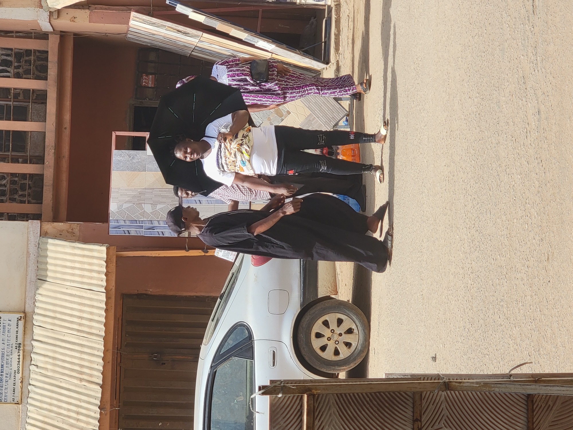 This outdoor street scene in Maiburiji, Nigeria, depicts several individuals and a white car under bright daylight. The image is taken from a slightly low angle, showing buildings on the left and a paved ground area.
In the center, a person wearing a white t-shirt with a colorful pattern and black trousers holds a large black umbrella over their head, looking towards the camera. To their right, a person in a purple and white patterned traditional outfit is visible. Behind the umbrella holder, another person in a checkered shirt is partially seen. Further left, a person dressed in a long black robe stands beside a white sedan car, facing away from the camera. The heads of two other individuals wearing dark caps are visible on the far left and right edges.
The setting appears to be a commercial area, with stacks of building materials, specifically tiles, leaning against a building in the background. Strong shadows on the ground indicate bright sunlight, and the umbrella suggests protection from the sun.
Notable details include a white sedan car, a black umbrella, and various building materials. On a white sign attached to a corrugated metal fence on the left, text reads: "ERS IN ALL KINDS OF BUILDING MATERINS S APV & RUVFERTE CERAMIC TILS, FICH CALL A FES VE, ELECT I ALR EUCTIONIC OF AL TYPES & GENERAL SUPPLIERS. AD. 3 KANO RD, KADUNA AD.2 KANO RD, KADUNA. 08036667098". A small red sign with the number "737" is also visible on a building. The overall scene captures a lively, sunny day in an urban environment.