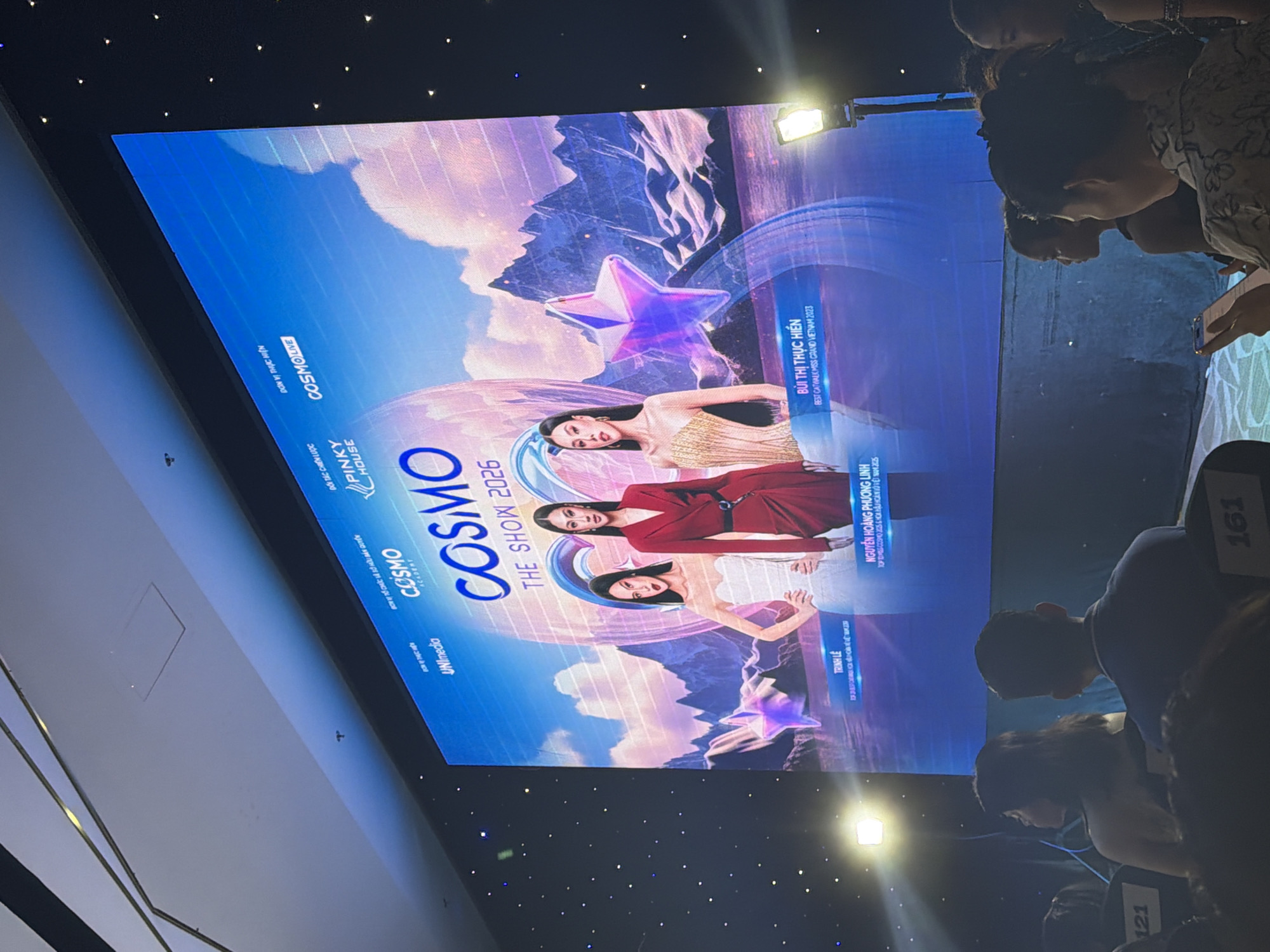 The image captures an indoor event, likely a beauty pageant or fashion show, held in a darkened venue in Nha Trang, Vietnam. The specific ward within Nha Trang is not discernible from the visual evidence.
The focal point of the scene is a large, brightly illuminated LED screen serving as a stage backdrop, displaying promotional graphics for "COSMO THE SHOW 2026". The screen features three elegantly dressed female models or contestants standing side-by-side, identified by text below them as Trinh Lê, Nguyễn Hoàng Phương Linh, and Bùi Thị Thực Hiện, along with their respective titles from 2023 beauty pageants (e.g., "BEST CATWALK MISS GRAND VIETNAM 2023" for Bùi Thị Thực Hiền). Various logos are visible at the top of the screen, including "UNImedia," "PINKY HOUSE," "COSMO LIVE," and "COSMO ACADEMY." The screen's background art depicts a stylized, fantastical landscape with snow-capped mountains, clouds, water, and luminous purple-pink star shapes.
In the foreground and midground, an audience is seated in rows of dark, auditorium-style chairs. Numerous individuals are visible from behind or in profile, primarily facing the screen. Seat numbers "121" and "161" are visible on the backs of chairs. On the far right, a woman in a floral-patterned shirt is seen looking down at a smartphone, while another woman next to her also looks towards the stage.
The venue's environment includes a dark wall on the right adorned with numerous small, scattered lights, creating a star-like effect. The ceiling in the upper left features modern architectural lines and what appear to be recessed light fixtures. Bright stage spotlights are positioned on either side of the stage, with a distinct light source creating a lens flare on the right side of the frame. The overall lighting is subdued, emphasizing the illuminated screen and stage.