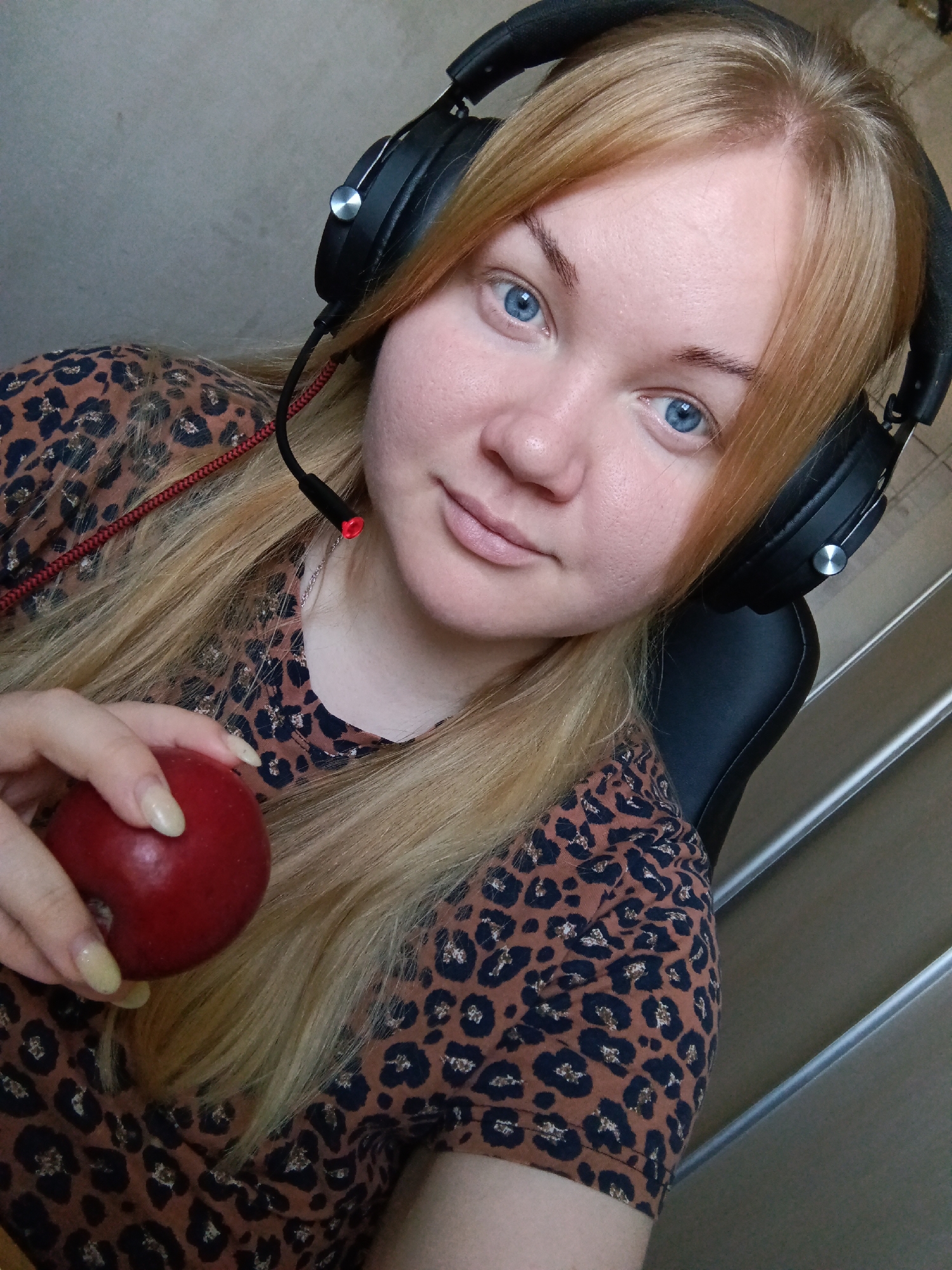A medium-shot, close-up, selfie-style image features a fair-skinned, blue-eyed woman with blonde hair, wearing black headphones with a red wire microphone, holding a red apple in her left hand. She is seated in a black chair and wearing a brown animal-print shirt. The background includes a plain wall and a staircase. No information is provided that would support any identification of a location, particularly in the city of Фоки, Russia.