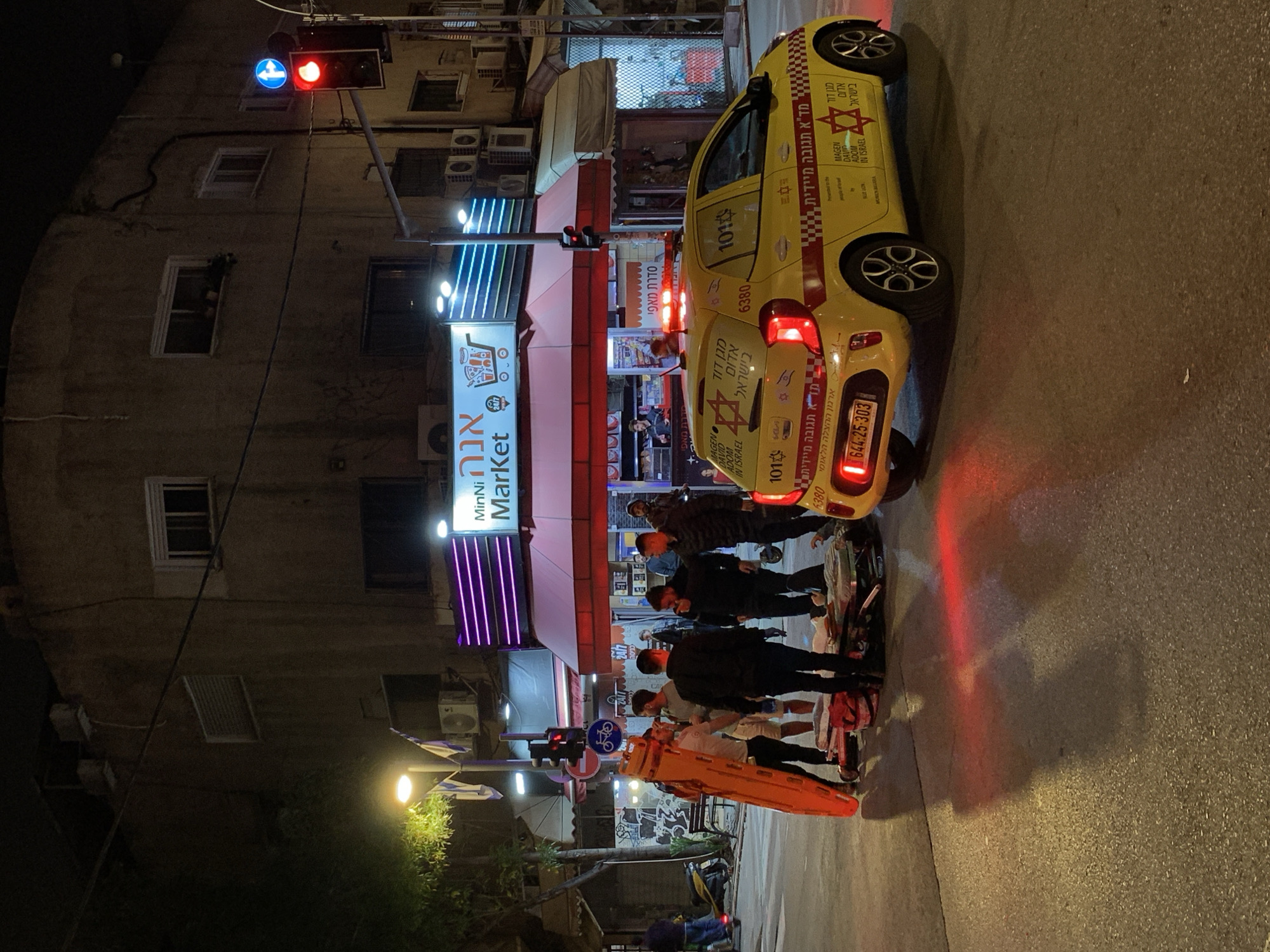 The image captures a street scene in Tel Aviv, Israel, at night. Prominently featured is a yellow ambulance affiliated with Magen David Adom (MDA), identifiable by the Star of David emblem and Hebrew text, parked on the right side of the street. The ambulance's license plate number is visible. A group of people gathers near the ambulance and along the street, some of whom appear to be emergency personnel based on the orange stretcher. There may be a medical incident occurring at the site.
On the left side of the street, there is a building and a storefront with a sign that says "Minni Market". The street is illuminated by streetlights and the storefront's neon lights. Traffic lights are visible. The environment seems to be a typical urban street with a mix of commercial and residential elements.