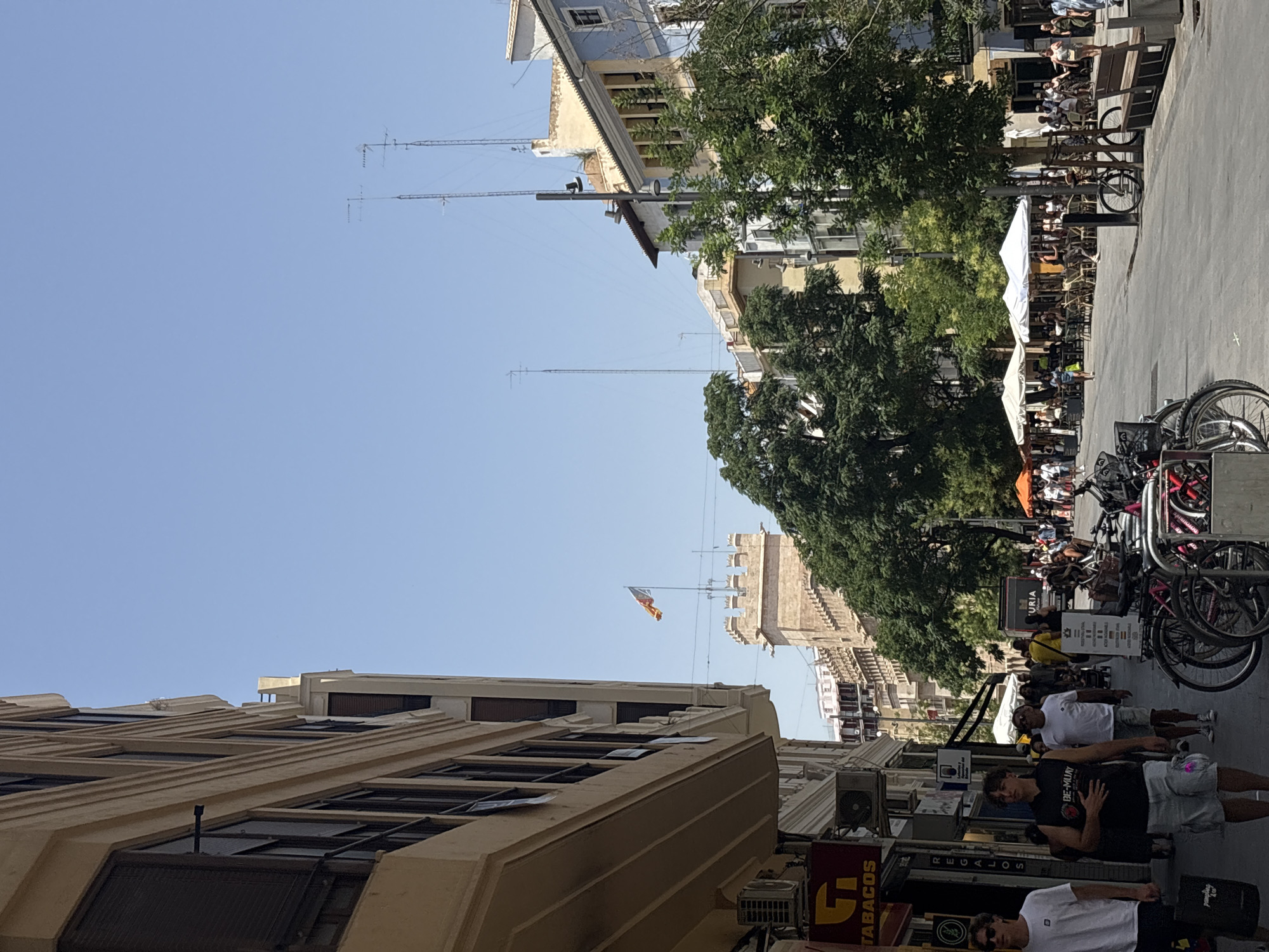The image depicts a street scene in Valencia, Spain, likely during the daytime on a clear, sunny day. The street is lined with buildings, some of which appear to be historic, with balconies and ornate architectural details. Trees are interspersed along the street, providing shade.
In the foreground, several people are walking along the sidewalk, dressed in casual summer attire. Bicycles are parked along the side of the street. In the background, there are outdoor seating areas for cafes or restaurants, with people seated at tables under umbrellas. A flag is visible on top of a tower in the distance. The overall atmosphere suggests a lively urban environment, possibly a commercial or tourist area.
