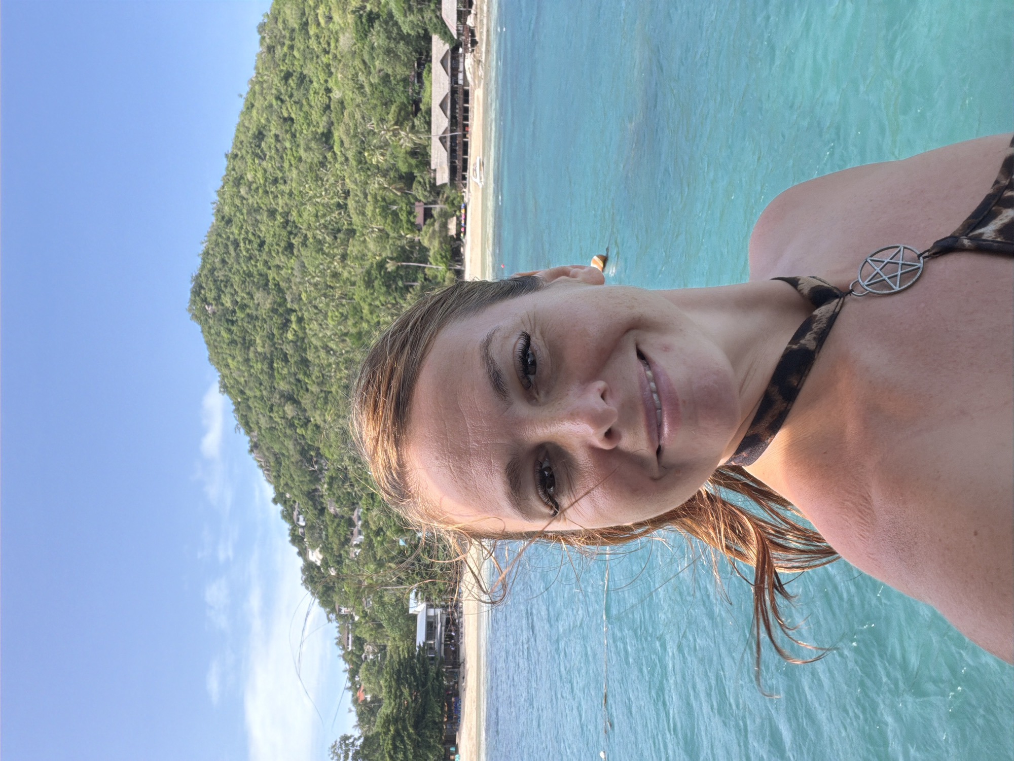 A woman is taking a selfie in the ocean in the Ko Pha-ngan District, Thailand. She is in the foreground, facing the camera with a slight smile. She has blonde hair that is windswept, and she is wearing a leopard-print bikini top with a pentagram necklace. The ocean is a vibrant turquoise color, and the background shows a lush green, tree-covered mountain and a beach with buildings. The sky is clear and blue with some scattered clouds.