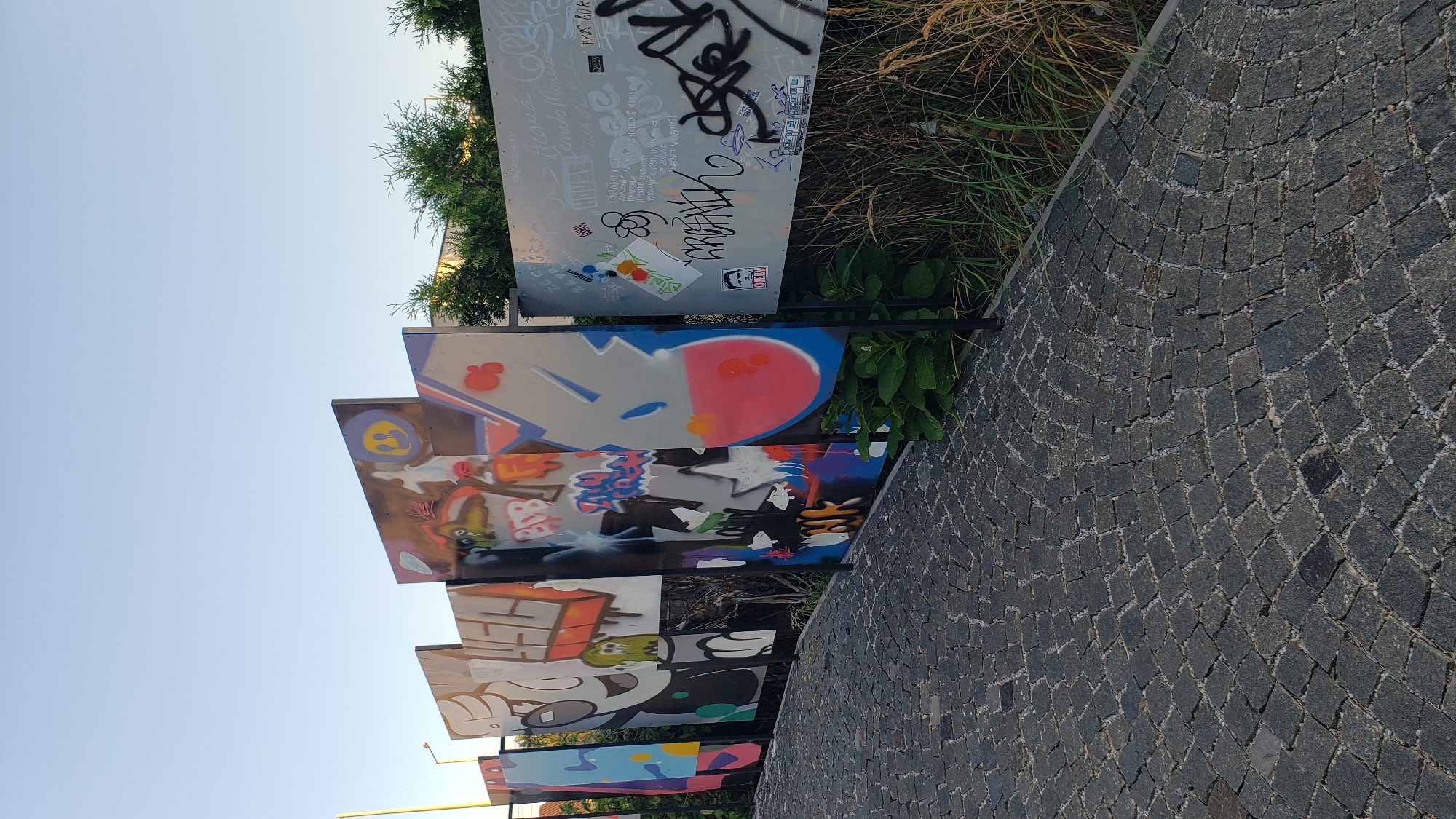 The image shows a section of the street art installation in Prešov, Slovakia, likely on or near Toryská street, given its proximity to the center and the artistic nature of the display. The foreground features a cobblestone pathway with a curving pattern, indicative of a pedestrian area or promenade. To the right of the path, dry grass and green foliage are visible, suggesting an outdoor, possibly park-like setting. A fence-like structure is lined with numerous rectangular panels displaying vibrant graffiti art. These panels are varied in size and orientation, creating a dynamic and layered visual effect. The artwork itself is diverse, featuring cartoonish characters, abstract shapes, bold lettering, and spray-painted designs in a wide array of colors. The background shows a clear blue sky and the tops of green trees, suggesting fair weather. The overall scene depicts a public space transformed by urban art.