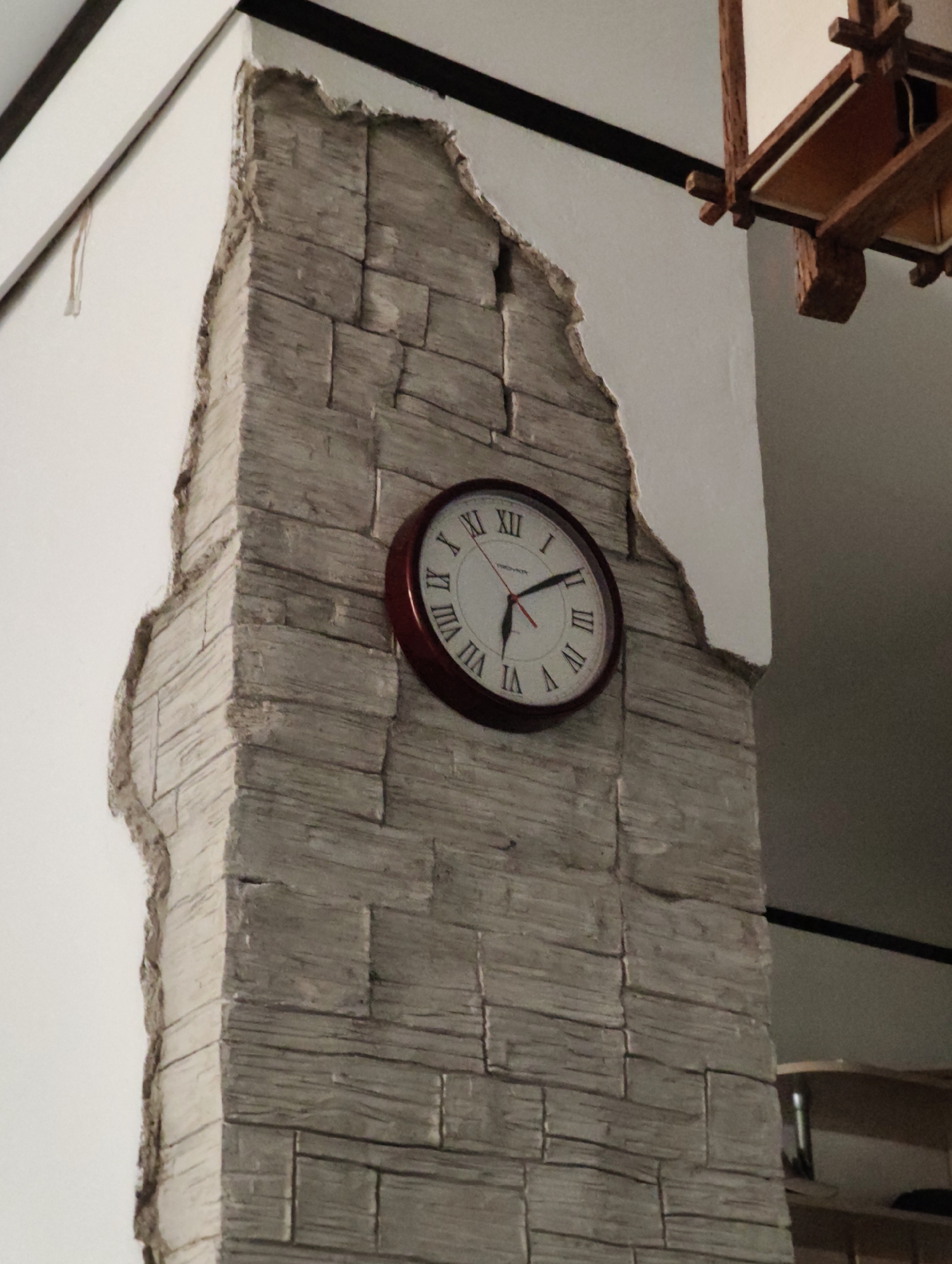 The image shows a close-up of a corner of a building with a decorative stone-like texture. A circular clock with Roman numerals and a dark red frame is mounted on the textured surface. The clock face is white with black hour markers and hands. The minute hand is pointing slightly past the 7, and the hour hand is pointing between the 10 and the 11, suggesting the time is approximately 10:37 or 10:38. The textured surface appears to be part of an interior wall or a decorative element on a pillar. The background is blurred, showing a hint of a ceiling with a wooden light fixture and a white wall. There is no indication of the city of Komsomolsk-on-Amur, Russia, or any specific location information within the image itself. The scene is static and appears to be a still photograph of an interior space.