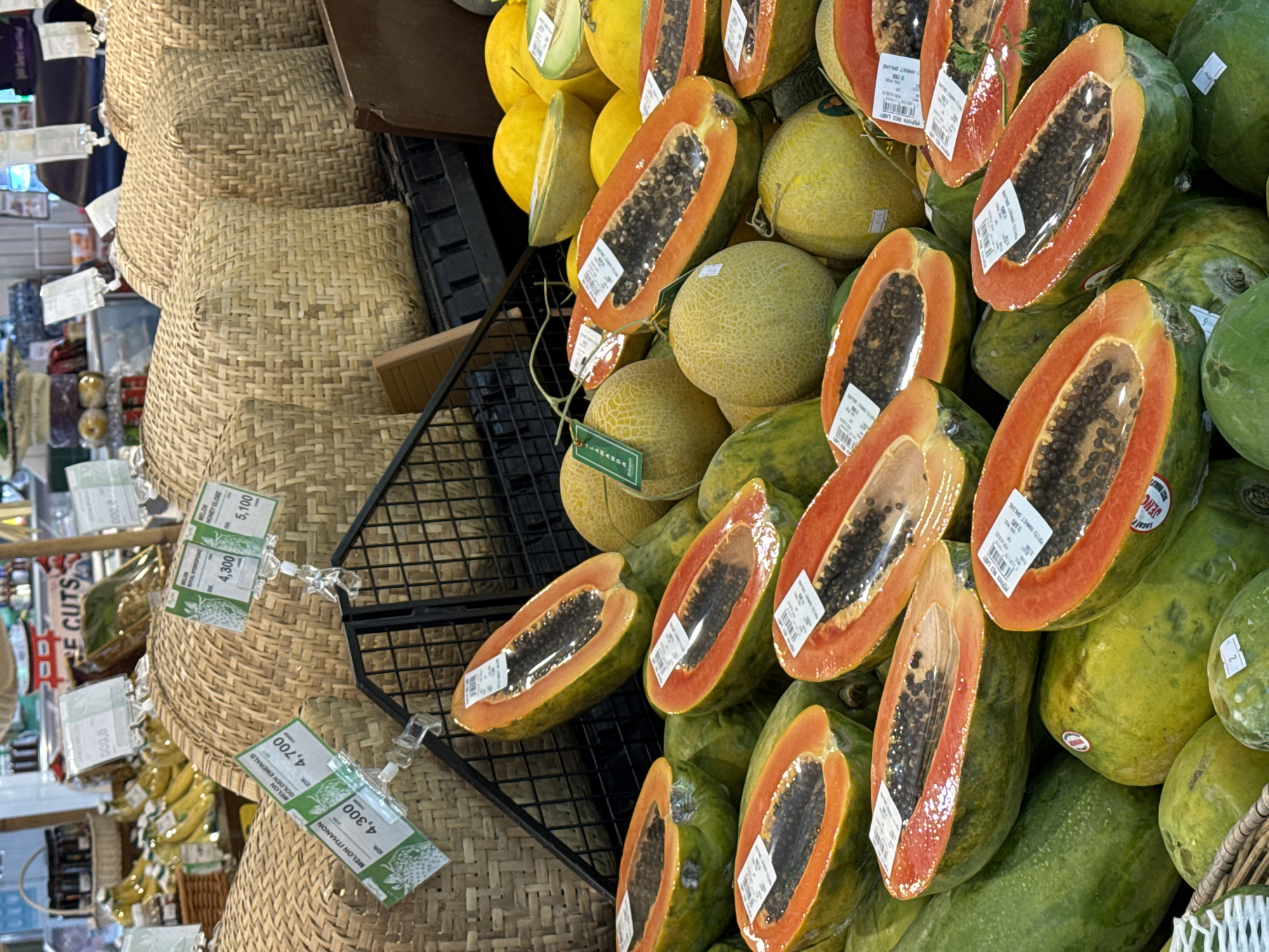 A high-angle, medium shot captures a fruit stall display in North Kuta, Indonesia, dominated by a vibrant assortment of papayas and melons. The papayas, predominantly cut in half lengthwise, reveal their bright orange flesh and numerous black seeds, each half individually wrapped in clear plastic. They are arranged in a dense cluster towards the foreground, with whole green papayas visible beneath them. Interspersed with the papayas are several whole melons, some with a netted rind and others smooth and yellow. The background features large, woven basket-like containers filled with more fruit. White price tags with green lettering are clipped to the baskets and some of the fruit. The overall environment appears to be an indoor market or grocery store, suggested by the ambient lighting and the presence of shelving and other produce in the blurred background.