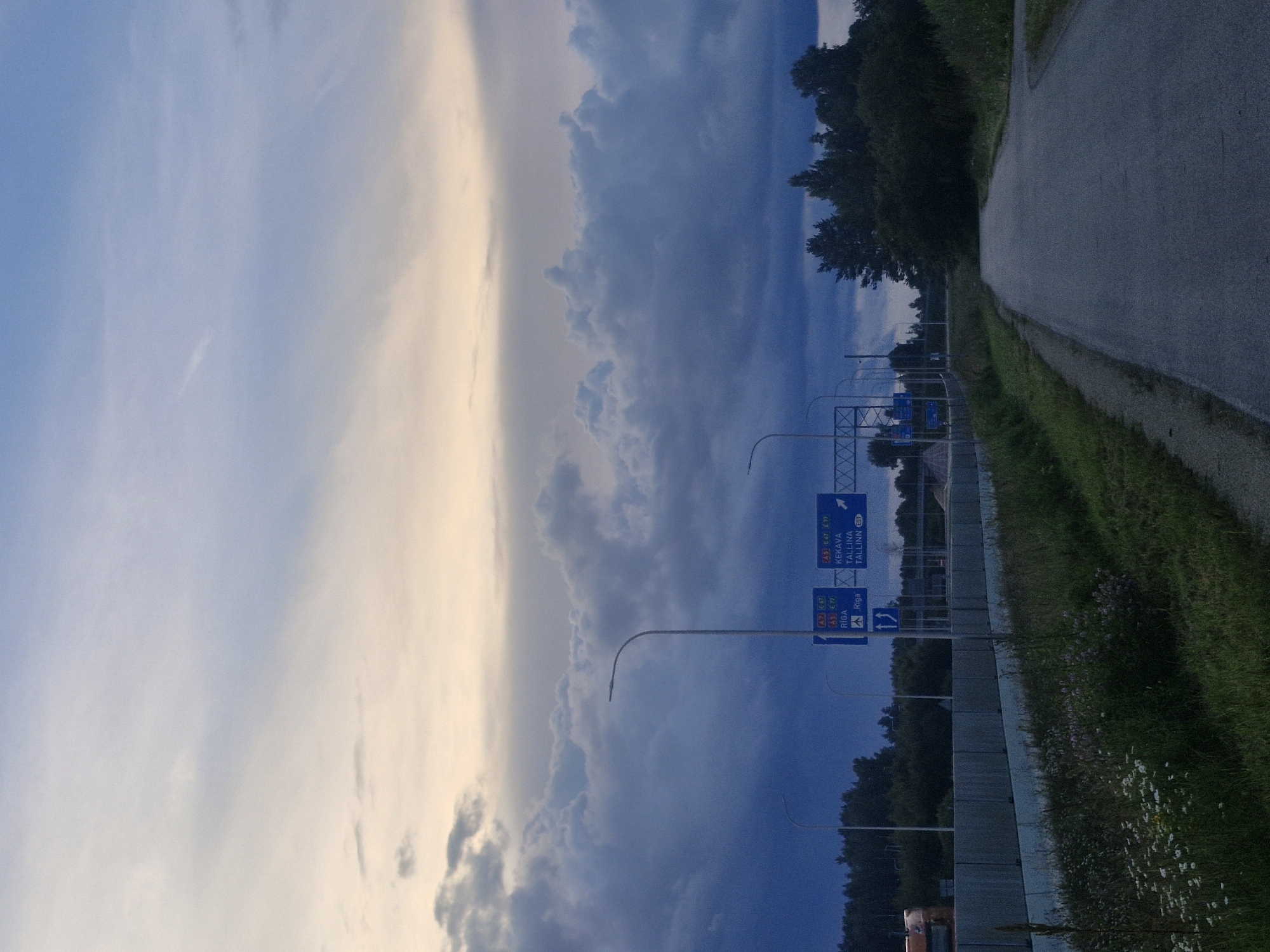 This image depicts a roadside scene in the vicinity of Kursīši, Latvia, under a dramatic, cloud-filled sky. The foreground shows a paved road with a grassy and flowered verge on the left, bordering a concrete barrier and a line of trees and bushes. On the right, the road continues into the distance, curving slightly to the right.
Above the road, a series of streetlights and directional signs are visible. One sign indicates "RIGA" with a distance of 140 KM and an airport symbol, accompanied by route numbers "A7" and "E67". Another sign points towards "KEKAVA" and "TALLINN" with route numbers "A5" and "E77".
In the background, the sky is dominated by dark, heavy clouds, with lighter, yellowish clouds forming a band near the horizon. The overall atmosphere suggests it might be dusk or a stormy weather condition. A glimpse of an orange truck is visible to the far left, behind the concrete barrier. The environment appears rural, with dense foliage and trees lining the far side of the road.