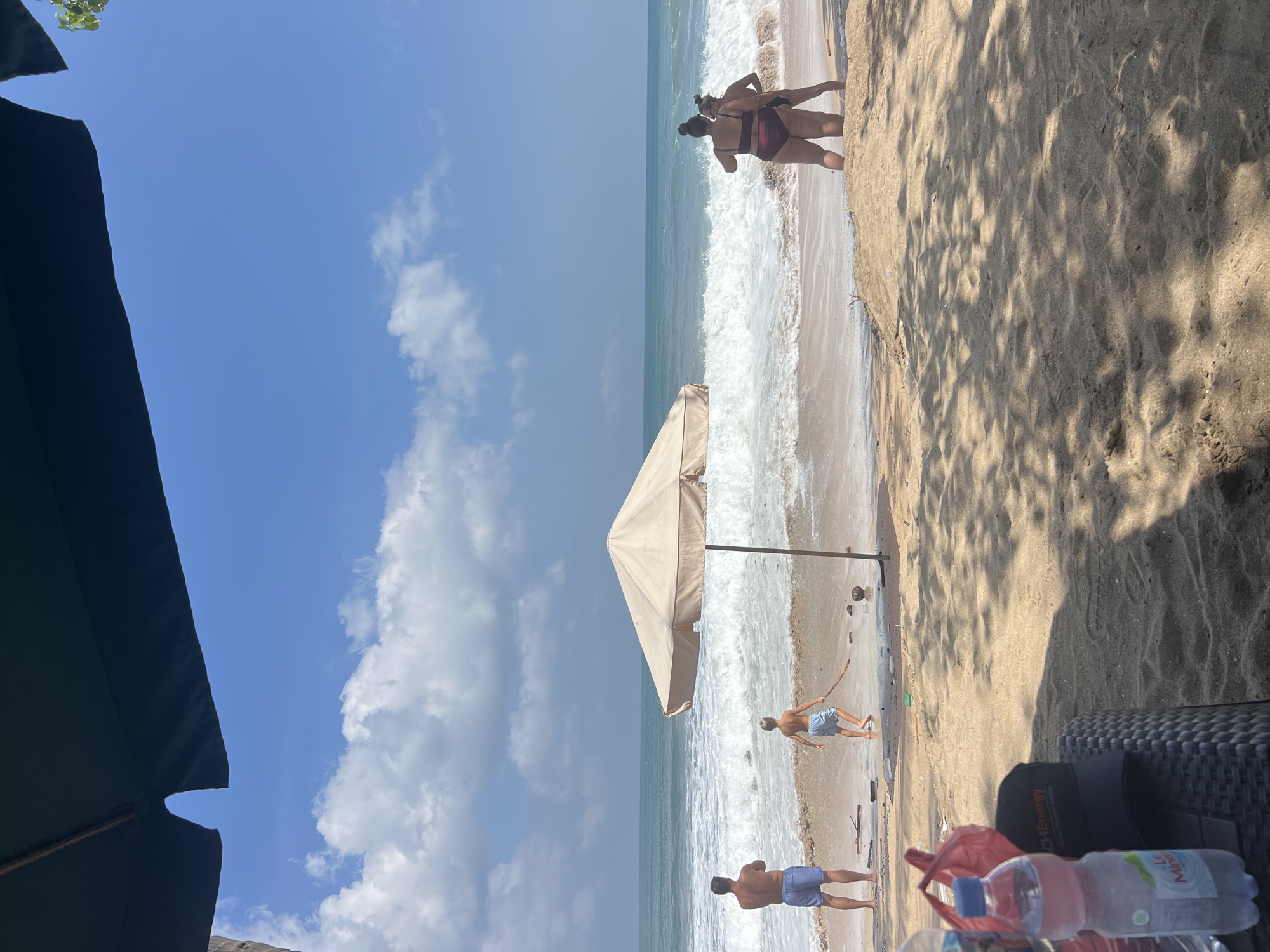 The image depicts a sunny day on Kuta Beach, Indonesia. In the foreground, sand is visible, with some shadows cast by overhead foliage. A woven table holds a plastic water bottle and a black baseball cap. Further back, a beige beach umbrella shades a section of the sand.
On the wet sand near the ocean, a man in blue shorts stands facing the sea. A young boy in blue swim trunks is also on the sand, facing away from the camera, holding a stick. Several pieces of driftwood and two coconuts are scattered on the beach. The ocean exhibits white-capped waves breaking on the shore.
On the right side of the frame, two women in bikinis are standing in the shallow water, facing away from the camera. The sky is blue with scattered white clouds. The overall scene captures a typical beach day with people enjoying the ocean and sand.