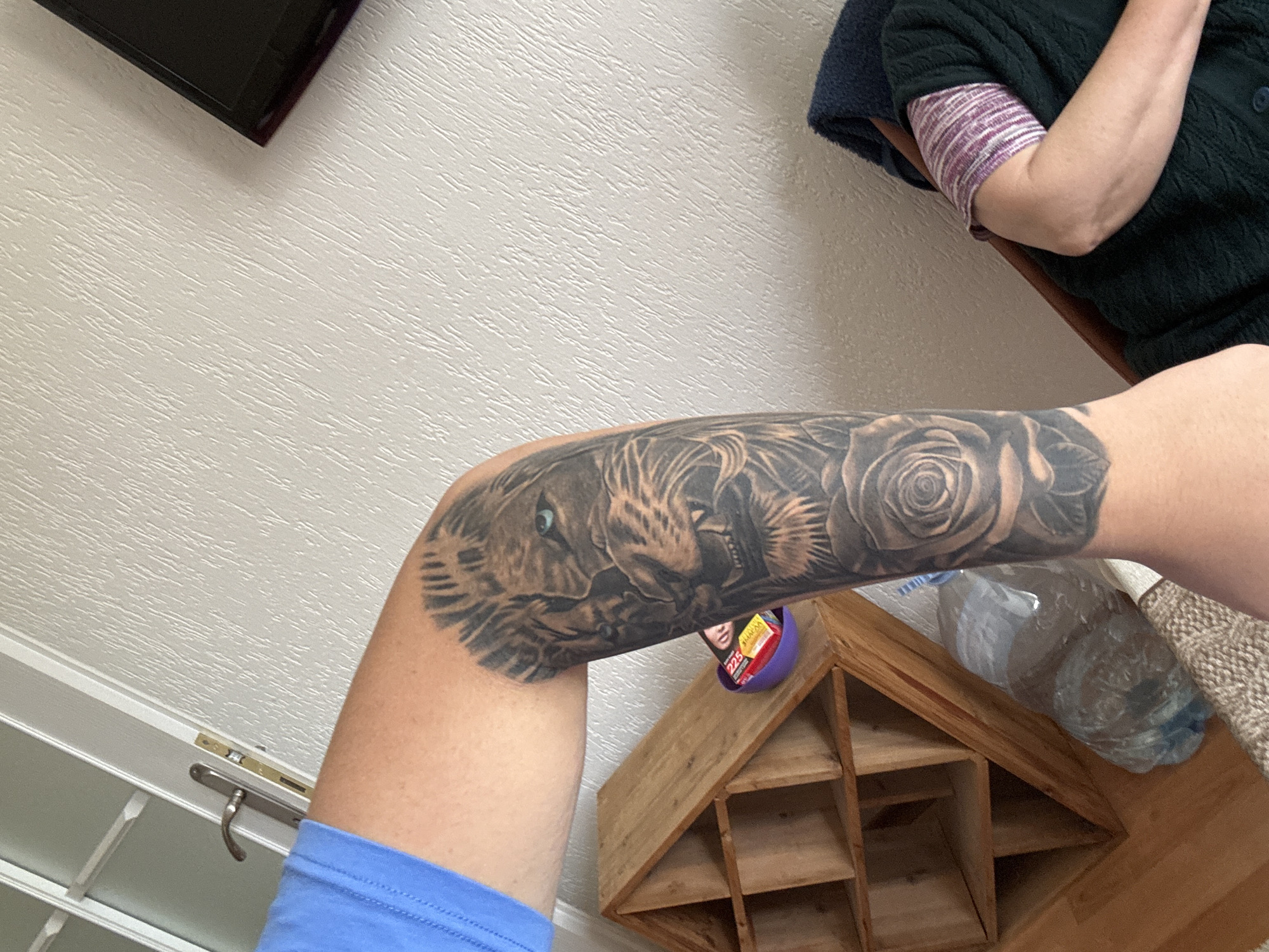 The image shows a person's tattooed forearm resting on a wooden shelving unit. The tattoo features a lion's head with one blue eye and a rose. A partially visible bottle of water and a purple container are also on the shelf. The person is wearing a blue garment. In the background, a television is mounted on a textured white wall. Another person with a dark green sweater is partially visible in the upper right corner of the image. The location appears to be inside a residence of Unknown, Unknown.