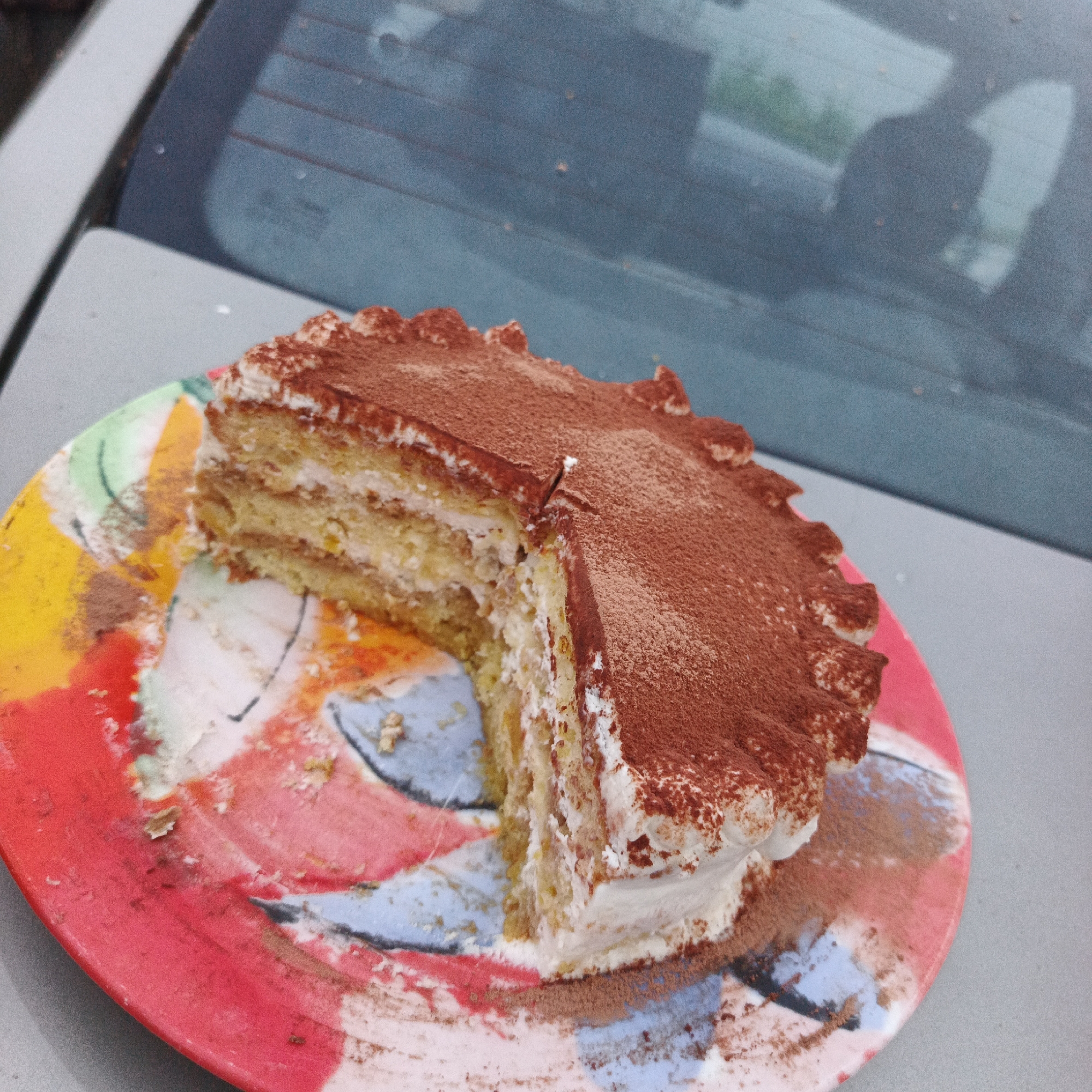 A sliced tiramisu cake is displayed on a colorful, abstract-patterned plate. The cake has layers of sponge cake, cream, and is dusted with cocoa powder on top. The plate is placed on a gray surface, possibly a car hood, with a blue, tinted reflection of a car window in the background. No people are visible in the image. The scene appears to be outdoors, with soft lighting. There is no indication of the location being Chaikovsky, Russia.