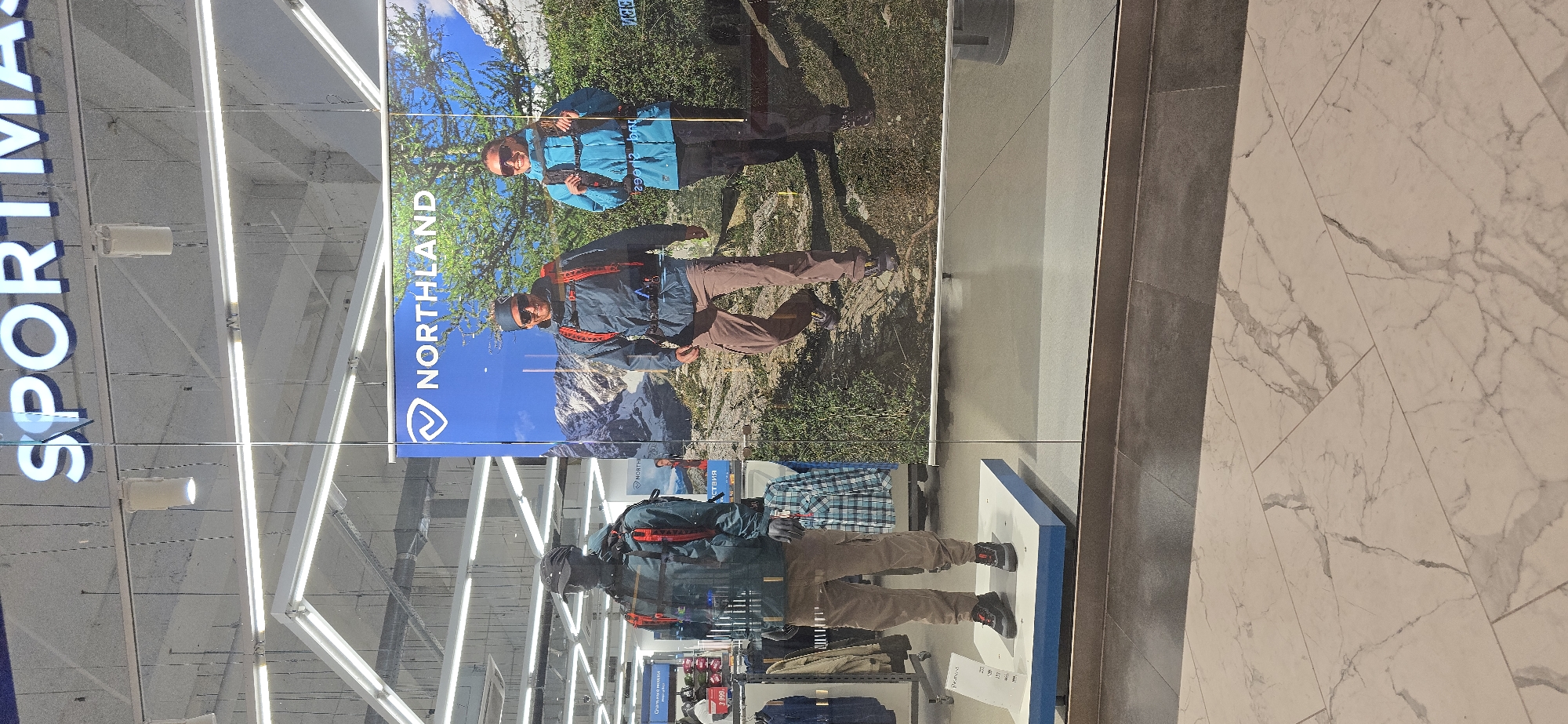 The image depicts the interior of a Sportmaster store. Visible in the foreground is a glass storefront displaying an advertisement for "NORTHLAND," featuring images of individuals in outdoor attire, presumably hiking or engaging in similar activities. Beneath the advertisement, a mannequin dressed in similar clothing stands on a platform. The store's interior extends into the background, showcasing clothing racks and merchandise. The location is likely in Grozny, Russia, based on the Cyrillic text on a nearby sign ("SPORTMASTER"). The foreground floor appears to be marble tile.