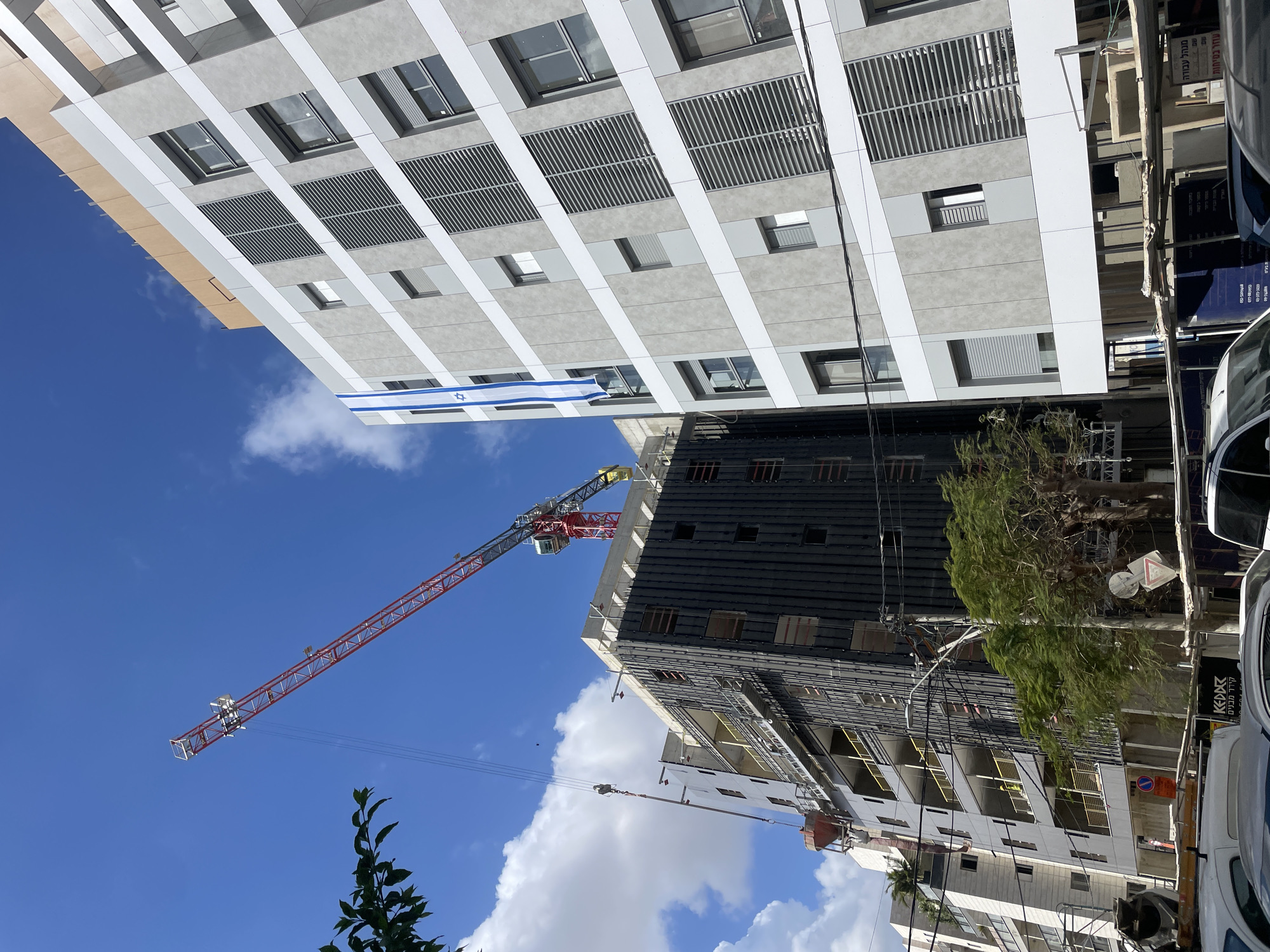 The image shows a construction site in Raanana, Israel. A tall, modern building is on the left, flying an Israeli flag. Adjacent to it is another building under construction, with a red and yellow crane towering above. Scaffolding is visible on the construction site, and a tree is positioned to the right of the building. Parked cars are visible in the right foreground. The sky is blue with scattered clouds.