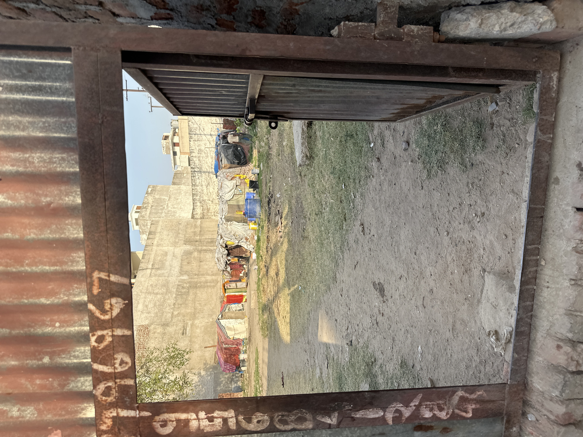 View through a rusty, metal frame, possibly a doorway or window, revealing an impoverished area in Mathura, India. The ground is dirt with sparse grass patches. Temporary shelters, constructed from various materials, line the edge of the open area, suggesting informal housing or a slum. A tall, unfinished concrete building forms the backdrop, with clotheslines and assorted discarded items visible. The scene suggests poverty and makeshift living conditions.