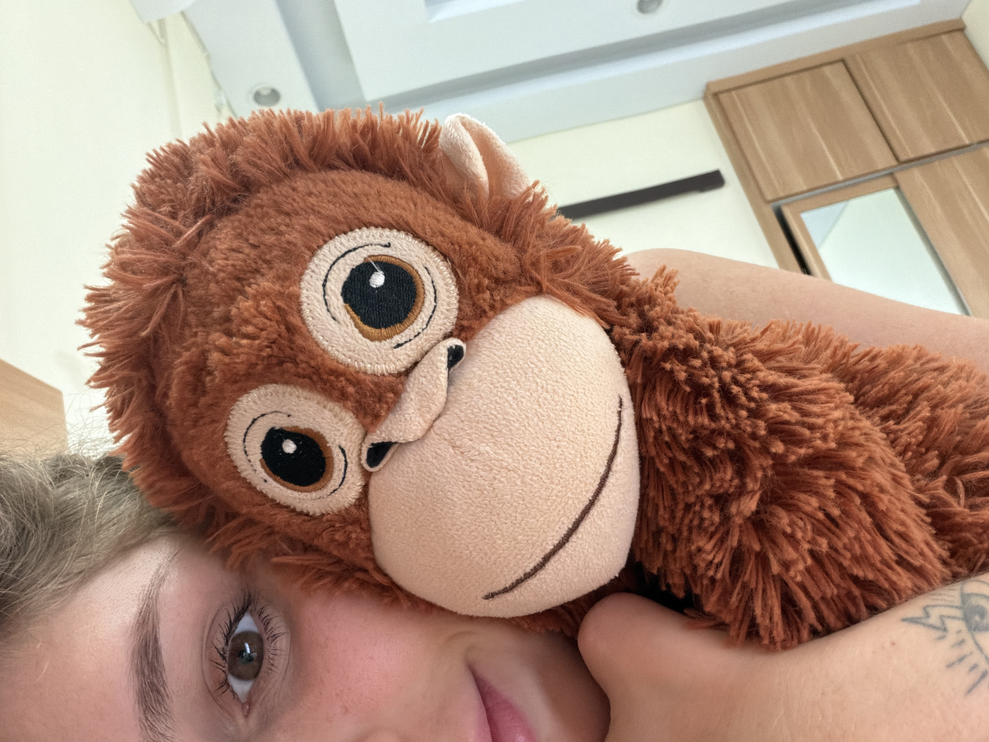 A close-up, high-angle photograph shows a person with light skin and brown hair lying down, with a brown plush monkey toy positioned against their head and upper body. The monkey toy has large, circular eyes with stitched details, a beige muzzle with a stitched smile, and brown fur. Part of the person's face is visible, including an eye, eyebrow, and part of their mouth. The person's right arm is also visible, with a tattoo on the forearm. The background includes a white ceiling with recessed lighting and a light-colored wooden wardrobe with sliding doors, one of which has a mirror. The location could potentially be in North Kuta, Indonesia, based on the appearance of the room and furniture, but this is not definitively verifiable from the image alone.
