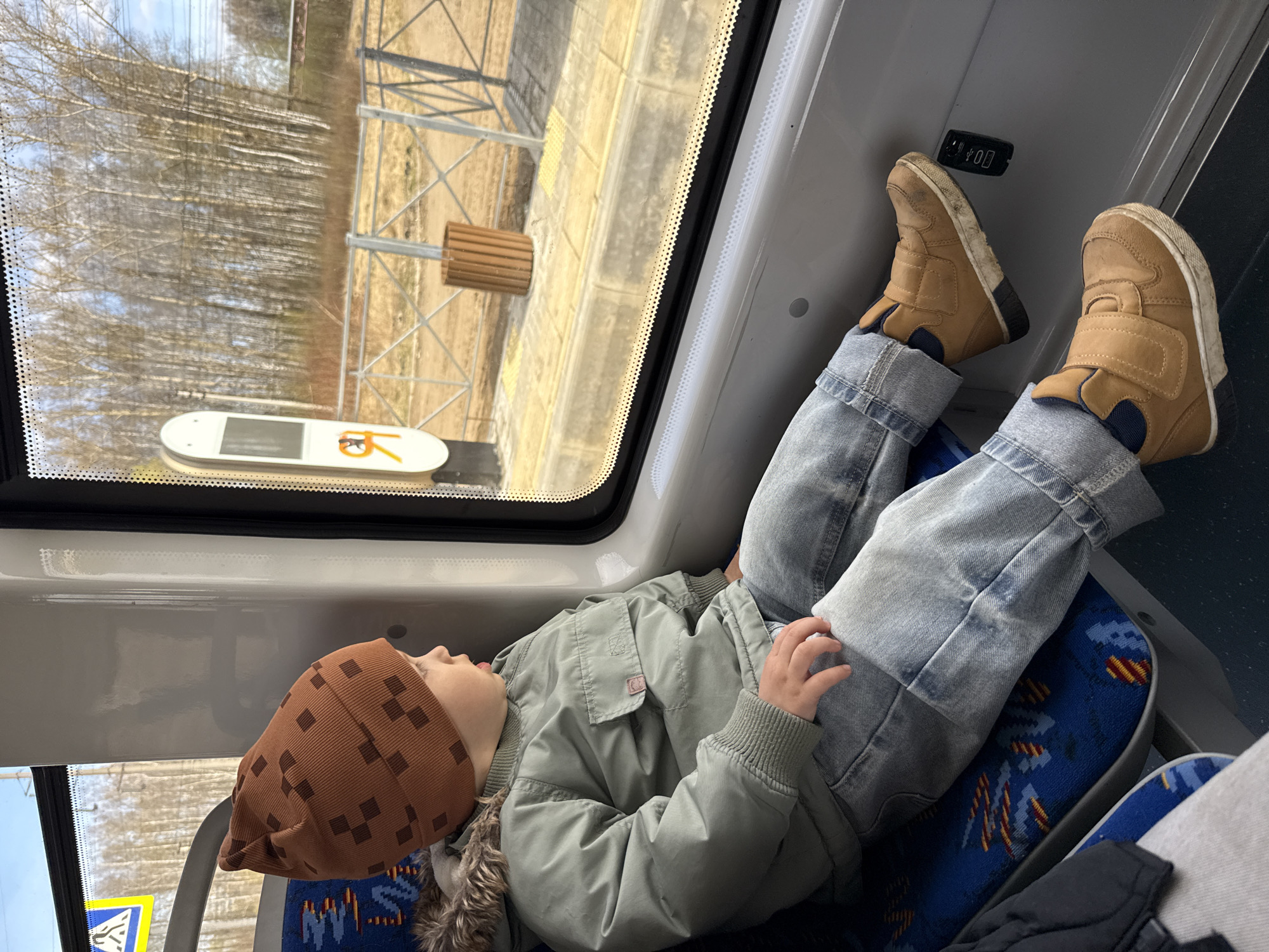 The image depicts a young child resting inside a train or electric commuter car in Rusanovo, Russia. The child, likely a boy, is seen lying on a blue-patterned seat, wearing a brown beanie with a repeating square pattern, a light olive-green jacket with a fur-lined hood, and light-wash blue jeans. His feet, clad in tan-colored high-top sneakers with Velcro straps, are propped up against the interior wall above a black USB charging port, which has two slots and faintly visible "USB" text. The child's mouth is slightly ajar, indicating he may be sleeping or deeply relaxed.
Through the large window, a daytime scene unfolds. Outside, a dense stand of slender, bare trees, resembling birch, stretches across the landscape, suggesting late winter or early spring. A light-colored ground surface, likely a train platform, is visible beneath the trees. On the platform, there are metal lattice structures and a wooden, slatted cylindrical object. A white rectangular sign with a dark screen and an orange symbol resembling "YK" is also visible outside the window. A partial view of a yellow and blue warning sign is present in the lower left corner of the interior. The image conveys a peaceful moment of travel.