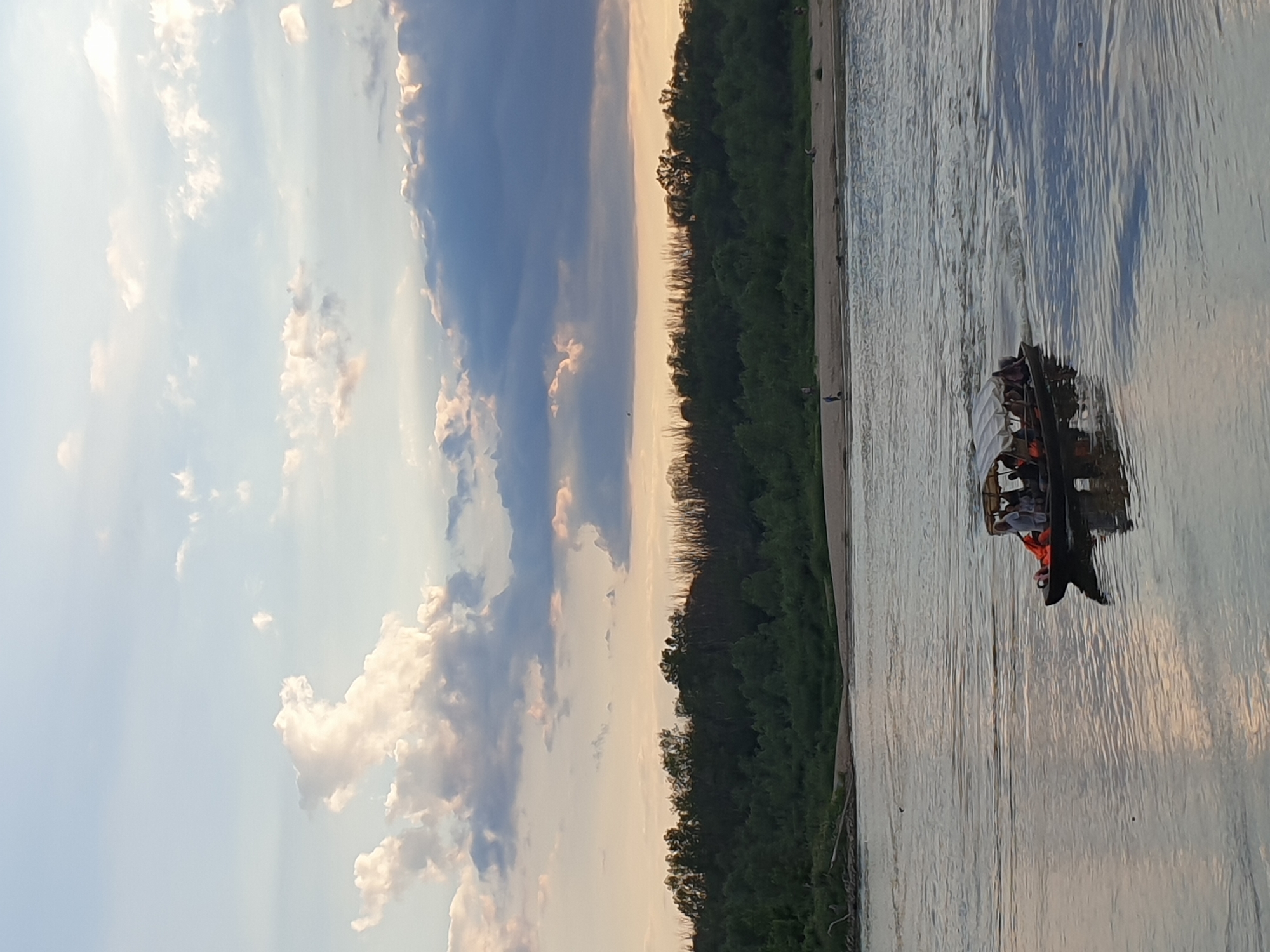 The image captures a wide river or lake scene at dusk, set in Unknown, Unknown. The upper portion of the frame features a sky transitioning from pale blue at the zenith to a soft orange-pink hue near the horizon. Scattered cumulus and stratocumulus clouds are visible, with their edges illuminated by the setting sun. A prominent, dense line of dark green trees and vegetation forms the far bank, bordered by a sandy shoreline. On this shoreline, several individuals are observed: one person walks on the left, accompanied by a small dark shape, while two other figures are discernible further to the right. In the mid-ground, a dark-hulled motorboat with a light-colored canopy moves across the water from right to left, leaving a distinct wake. Approximately five to six people are aboard the boat; one individual is prone in the bow, wearing an orange life vest, another person stands centrally in a white shirt, and additional passengers are seated under the canopy. The calm water surface reflects the muted colors of the sky and the boat itself. The overall lighting suggests late afternoon or early evening.