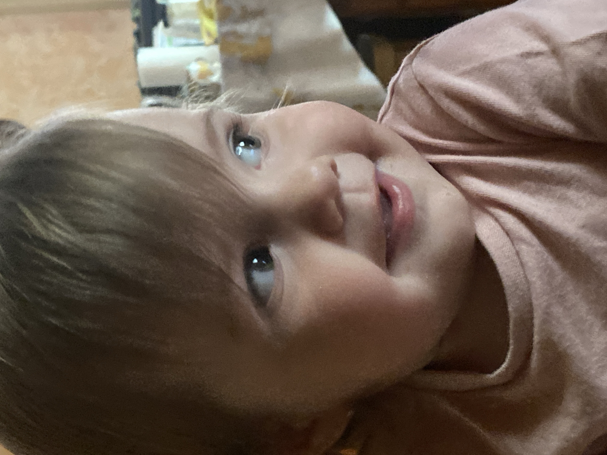 The image is a close-up, low-angle, portrait-style photograph of a young child lying down. The child has light brown hair and is wearing a pale pink shirt. The child's face is turned upwards, looking towards the camera with a slight smile. Their right eye is clearly visible, reflecting light, and their mouth is slightly open, showing the tip of their tongue. The lighting in the image suggests an indoor setting, possibly with natural light coming from above. The background is out of focus but appears to show some indoor furniture and possibly objects on a surface. The overall mood is warm and intimate, capturing a candid moment with the child. There is no visible text in the image.